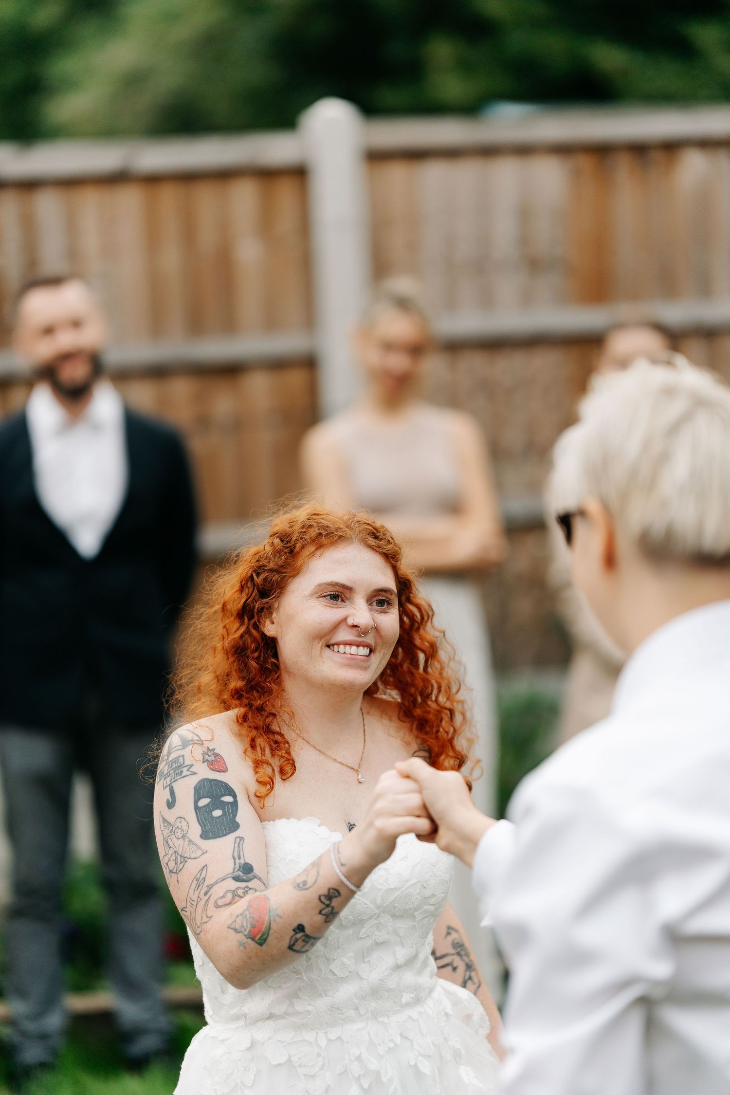 Tattooed redhead bride smiles while holding hands with partner during intimate backyard wedding ceremony.