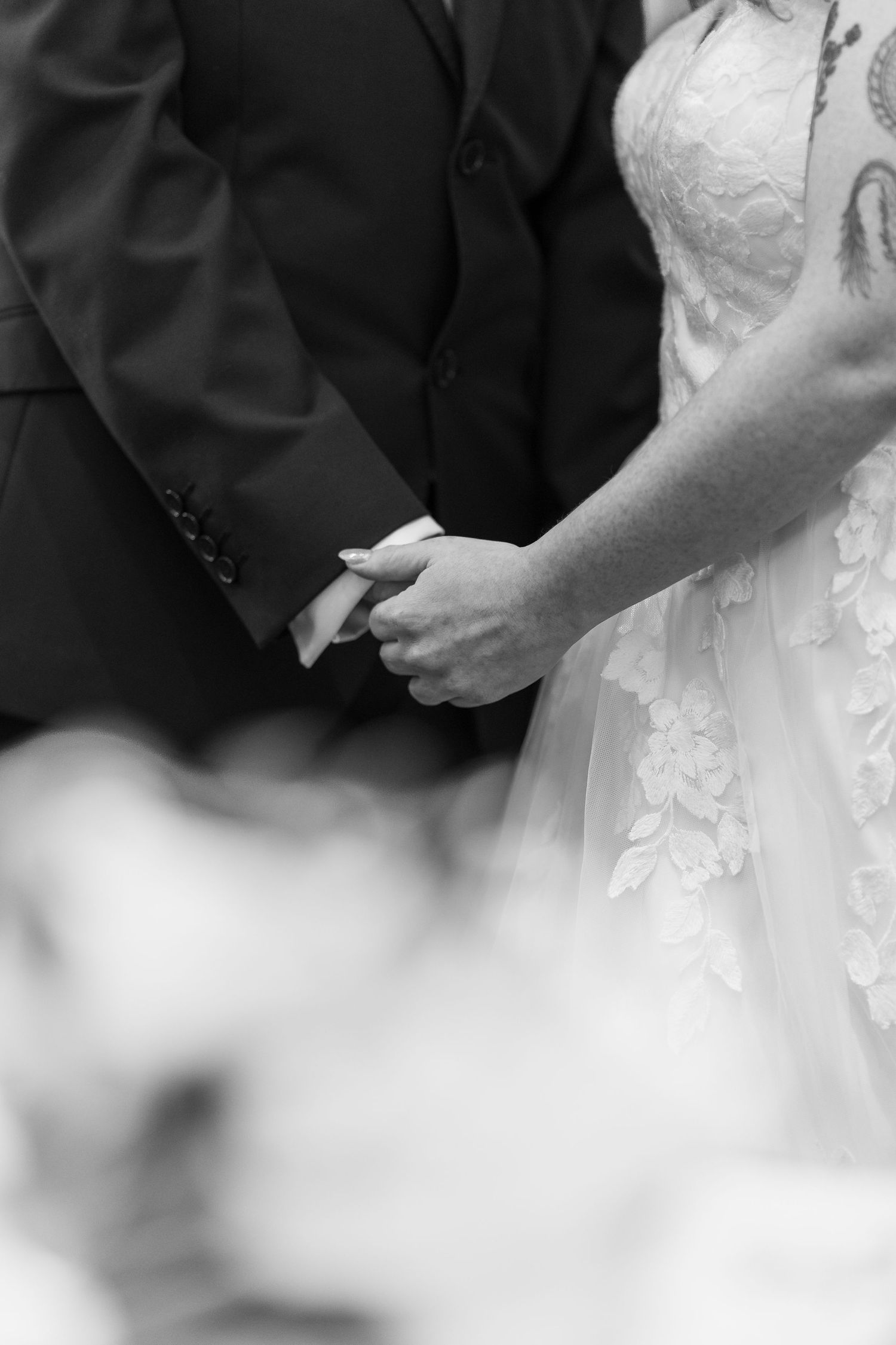 Black and white photo of bride and groom holding hands during wedding ceremony, bride in lace gown.