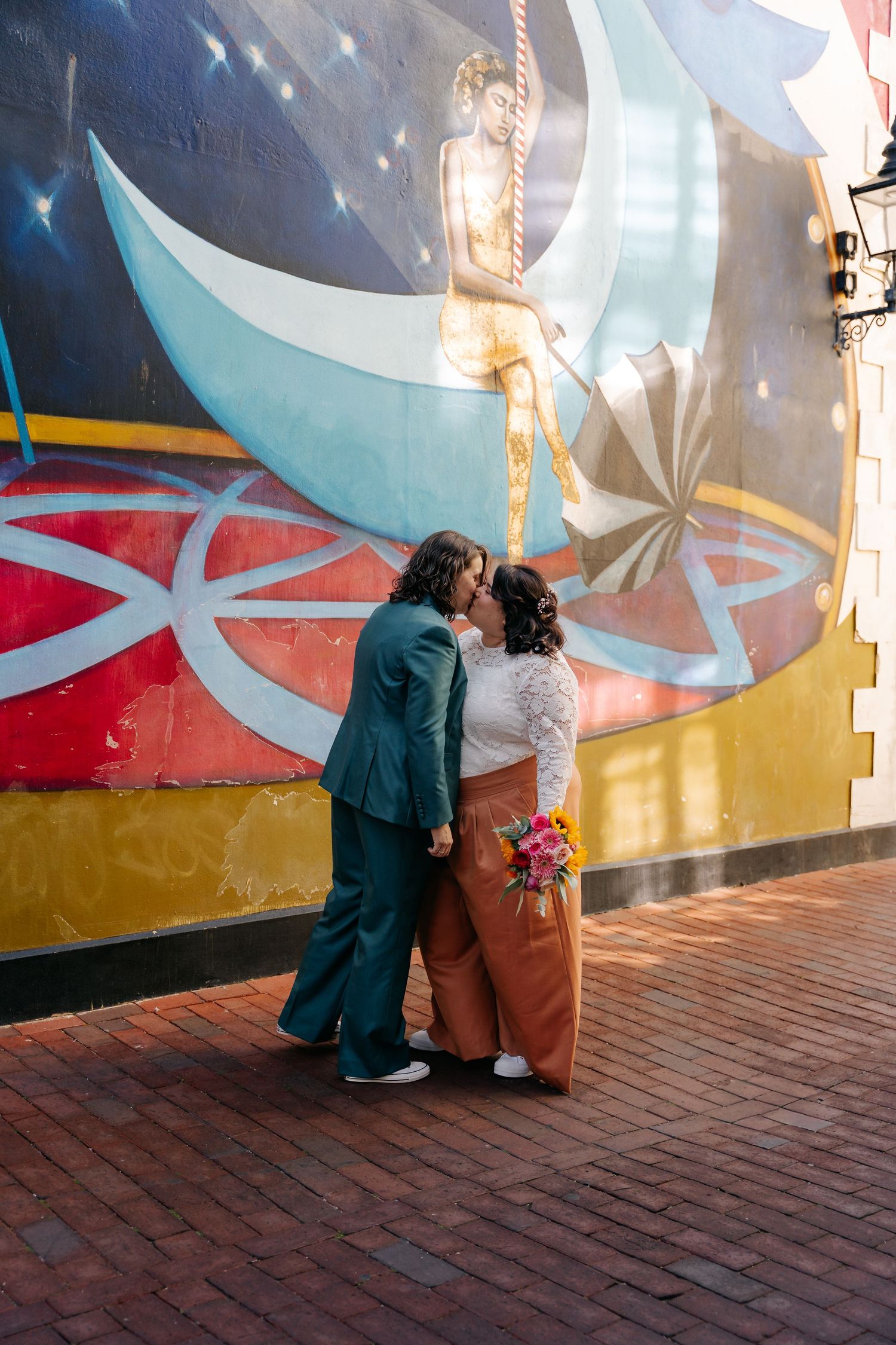 Two brides share a kiss in front of a colorful mural, one in a teal suit and one in rust pants with a floral bouquet.