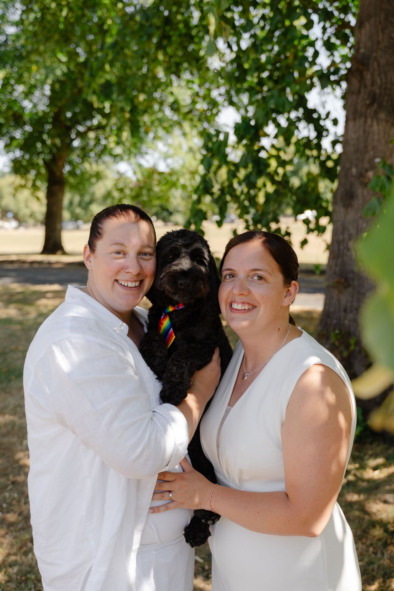 Two brides in white holding a black dog with a rainbow collar, smiling outdoors under green trees.
