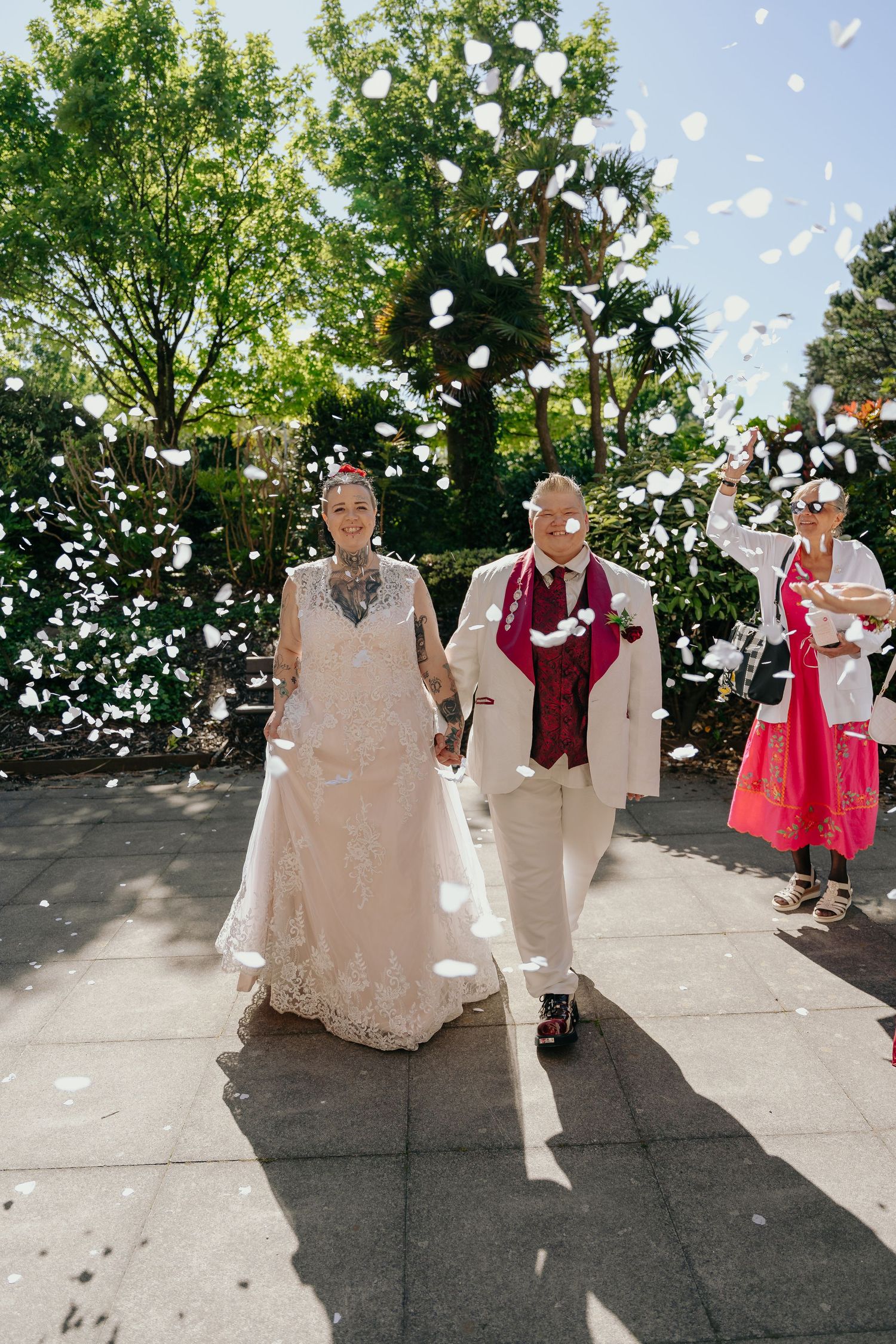 Bride and groom walk hand in hand through white confetti shower outdoors on sunny wedding day.