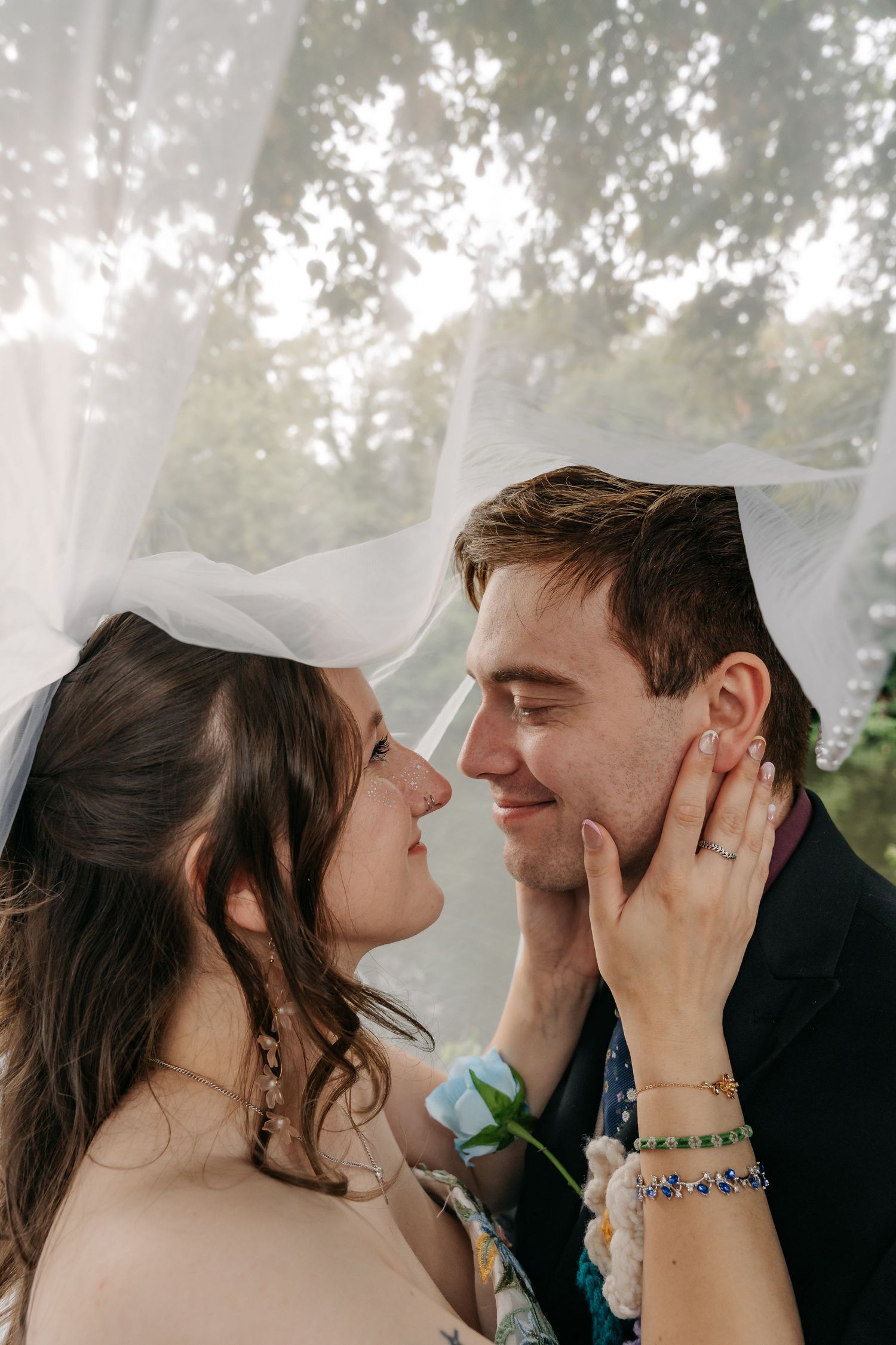 Bride and groom share a romantic moment under a flowing veil, with the groom smiling as the bride gently cups his face.