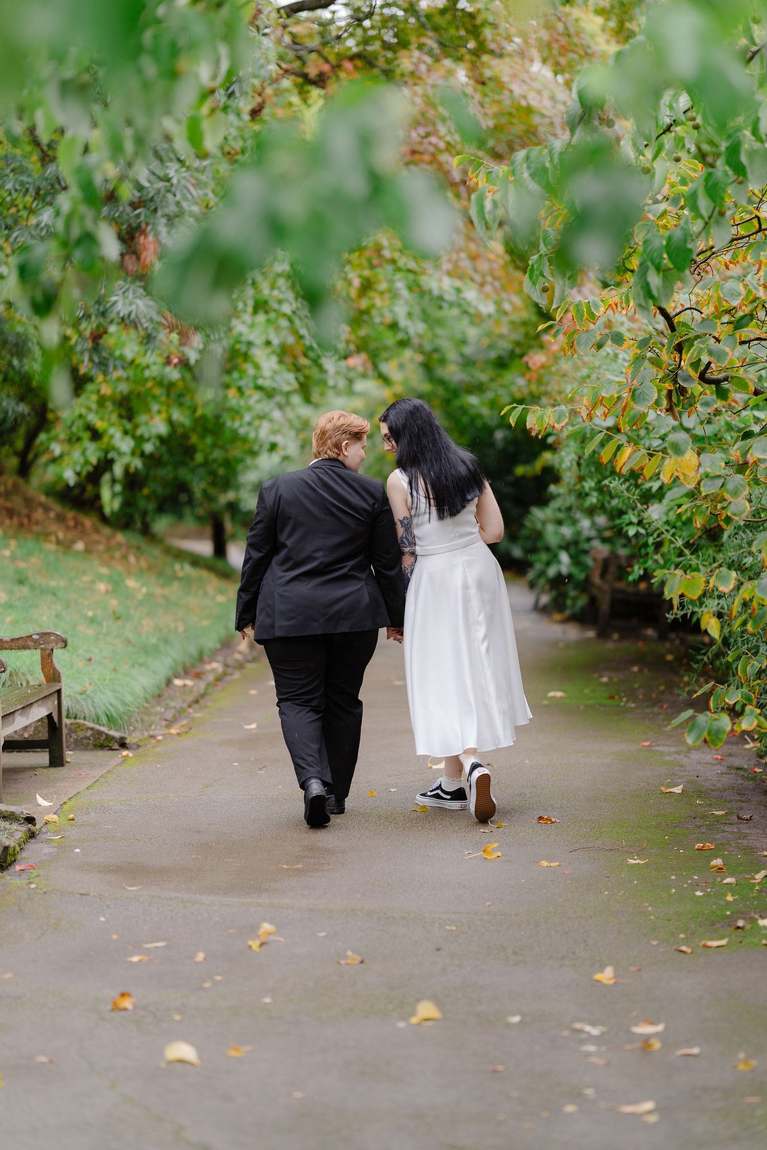 Two brides walk hand-in-hand down a tree-lined path, one in a black suit and one in a white dress.