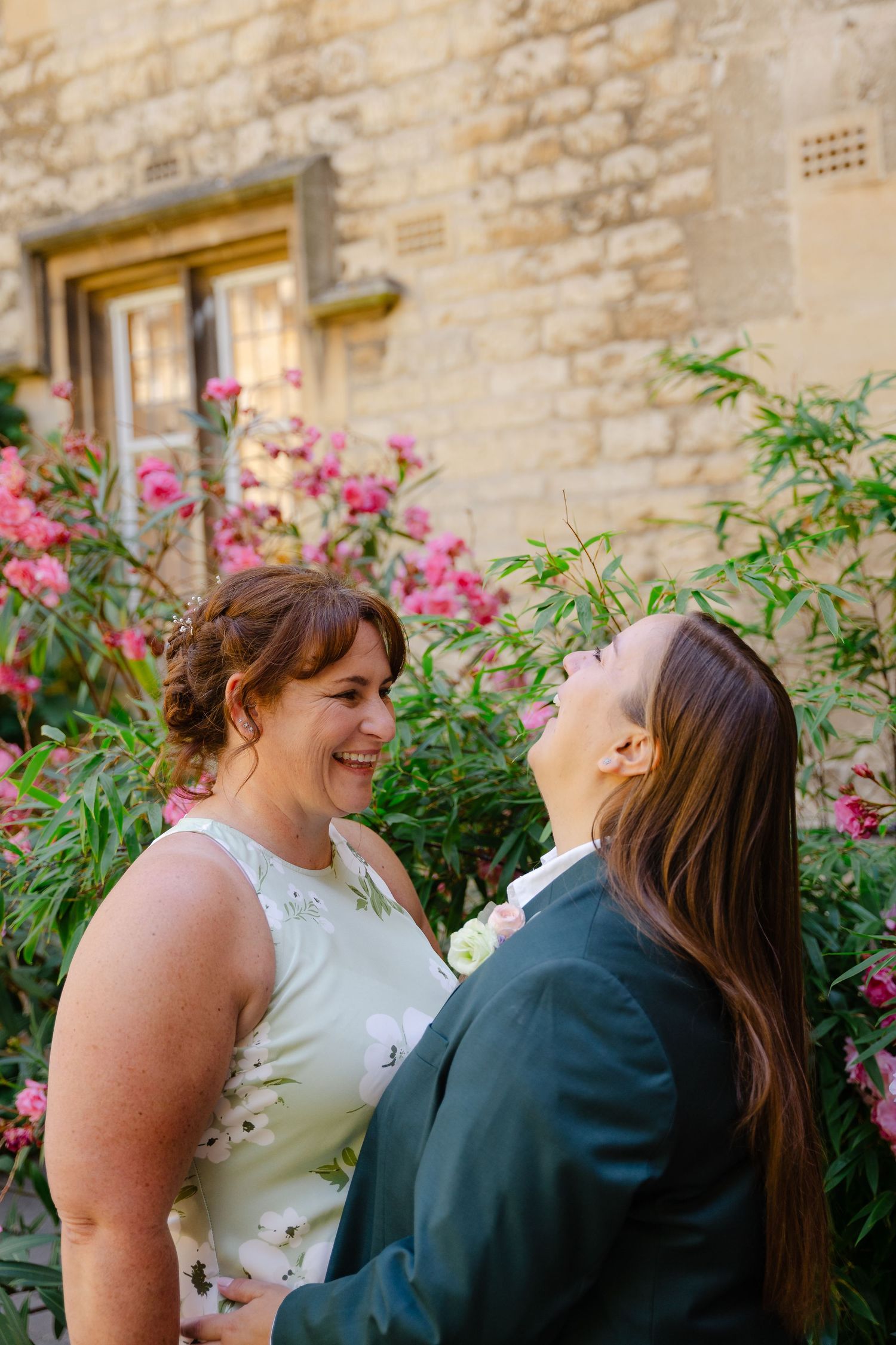 Two brides share a joyful moment outdoors, surrounded by pink flowers and a stone building backdrop.