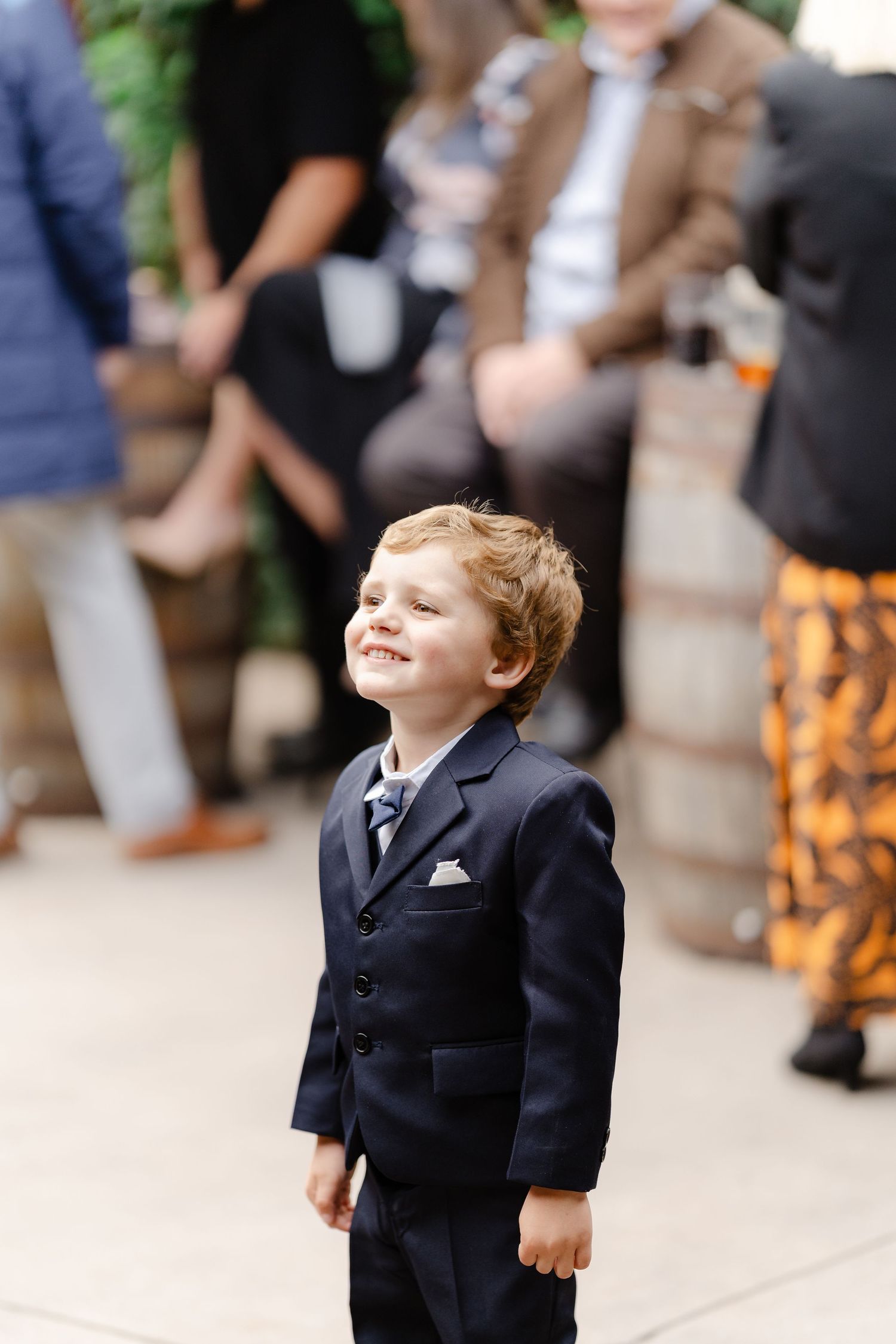 A smiling young boy in a navy blue suit with a pocket square stands happily at a wedding event.