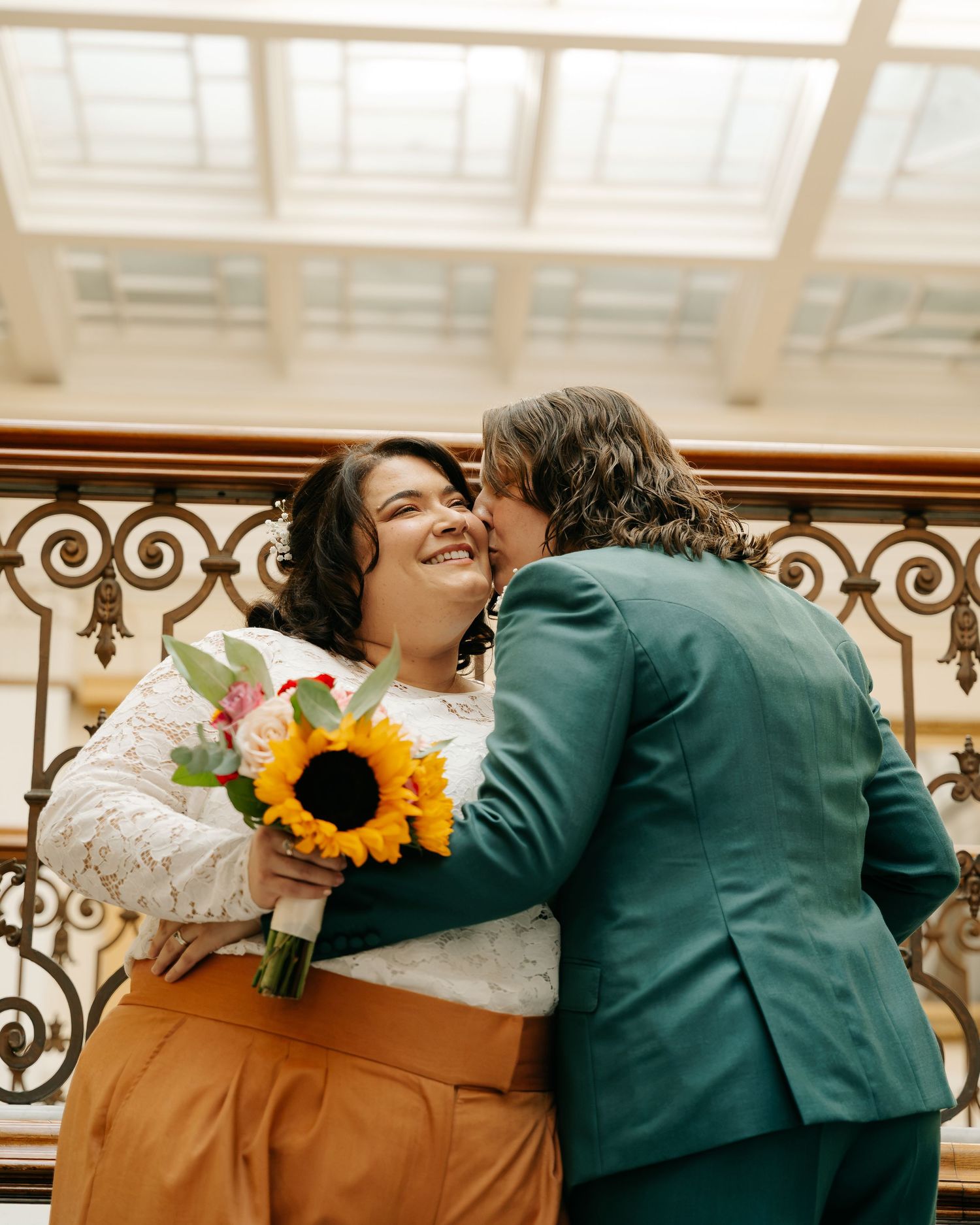 Two brides embrace on ornate staircase, one holding sunflower bouquet, wearing teal suit and lace dress.