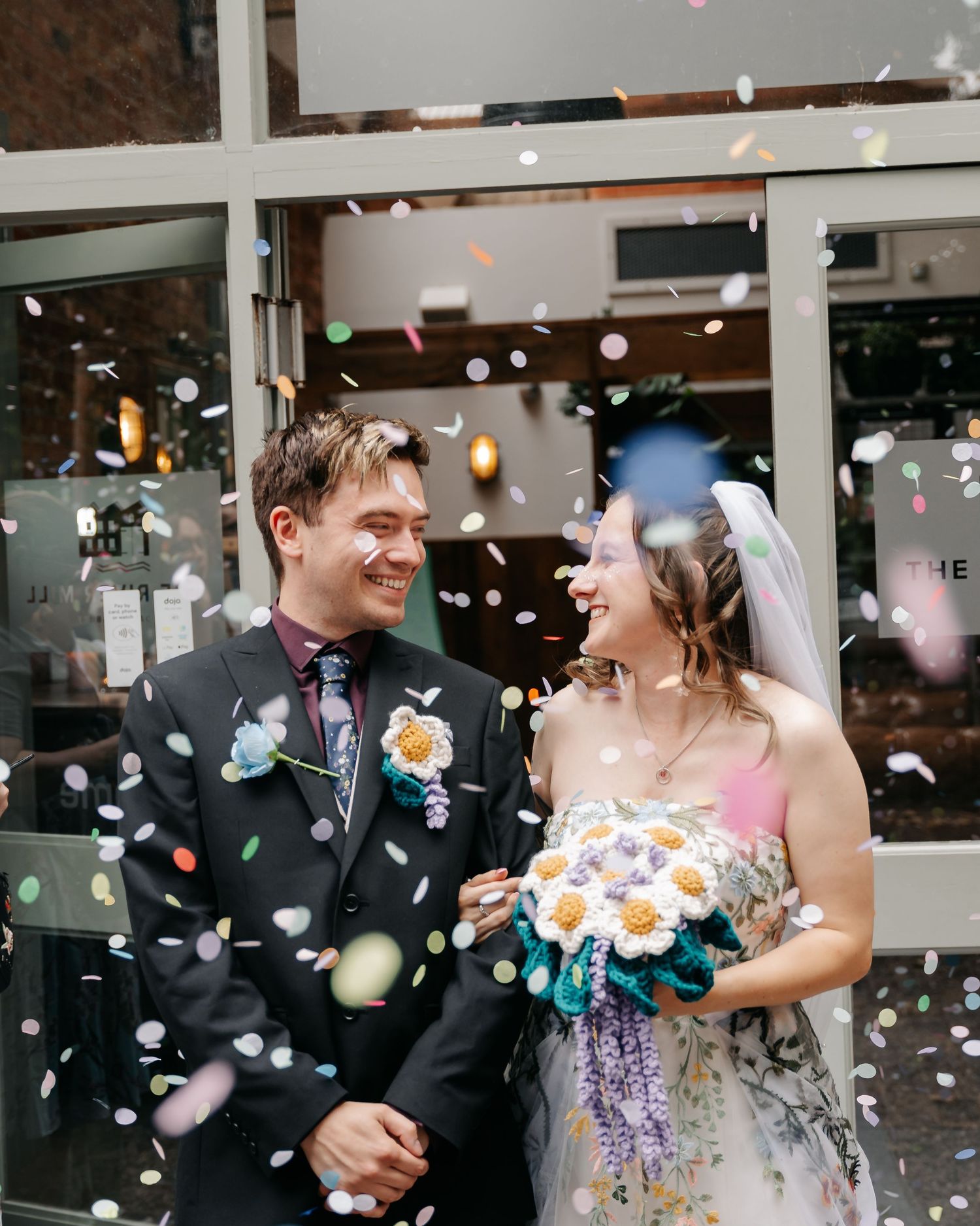 Happy couple laughing in confetti shower outside wedding venue, bride holding floral bouquet in floral gown with veil.