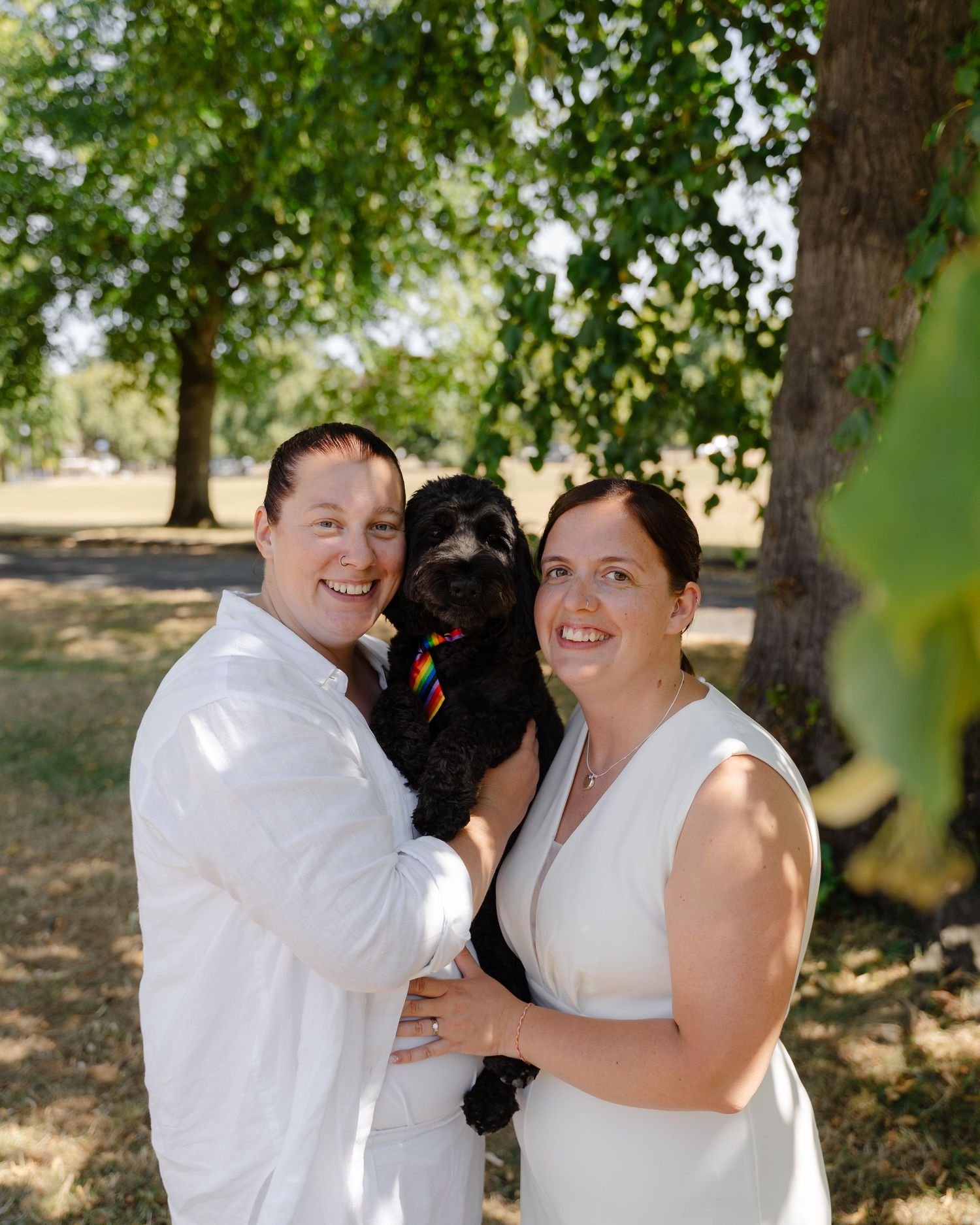 Two brides in white holding a black dog with a rainbow collar, smiling outdoors under green trees on a sunny day.
