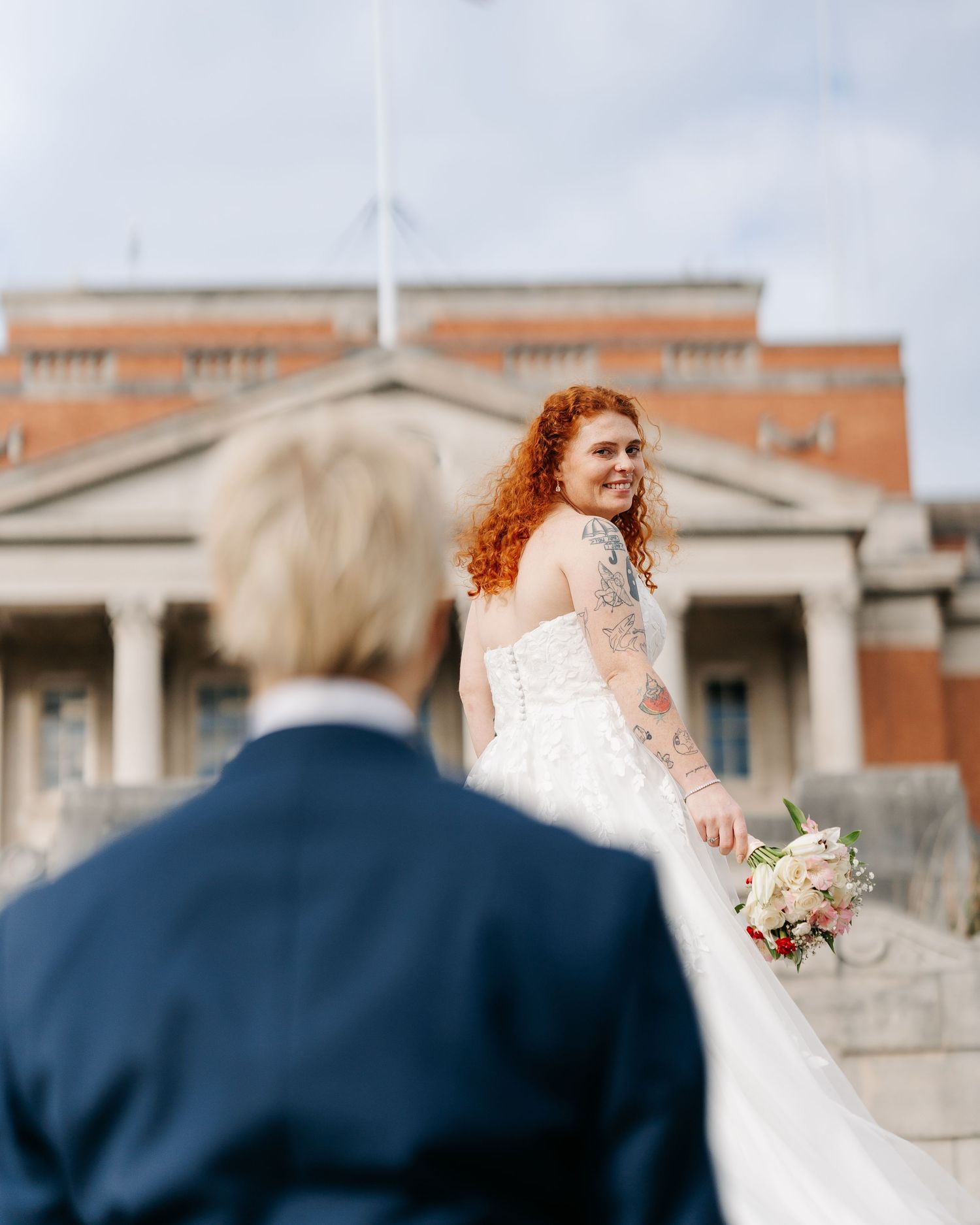A redhead bride with tattoos holds a bouquet on steps, smiling back at a person in a navy suit outside a brick building.