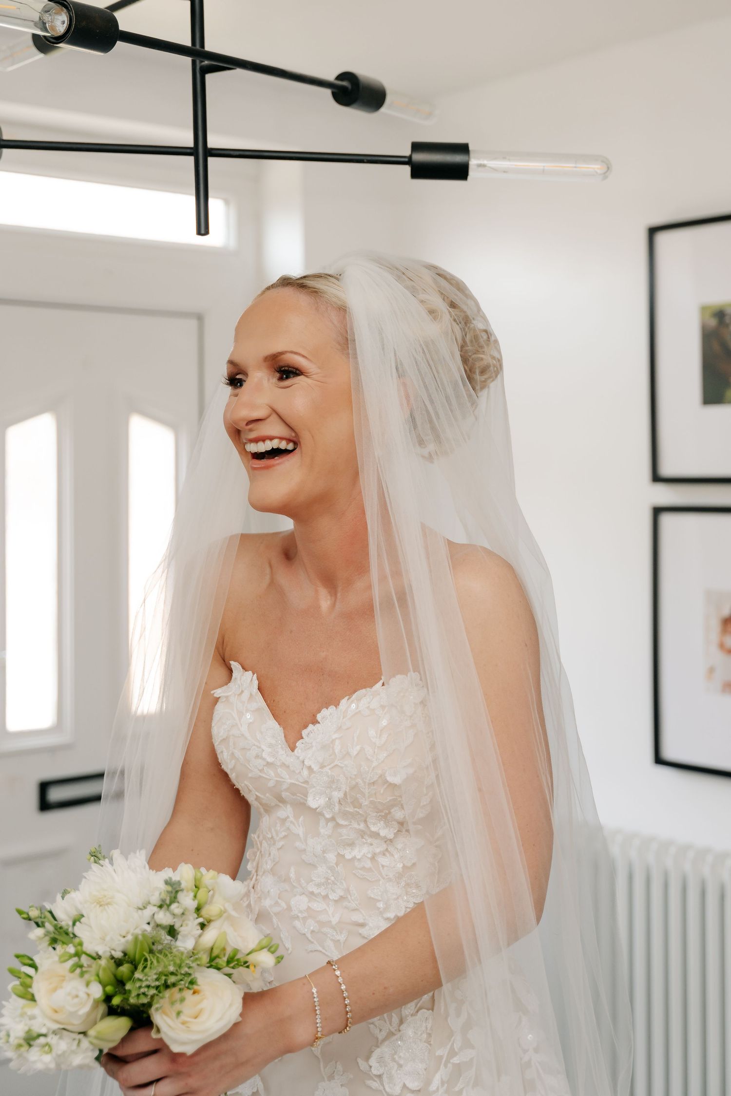 Smiling bride in a lace strapless wedding dress and veil holds a white bouquet indoors.