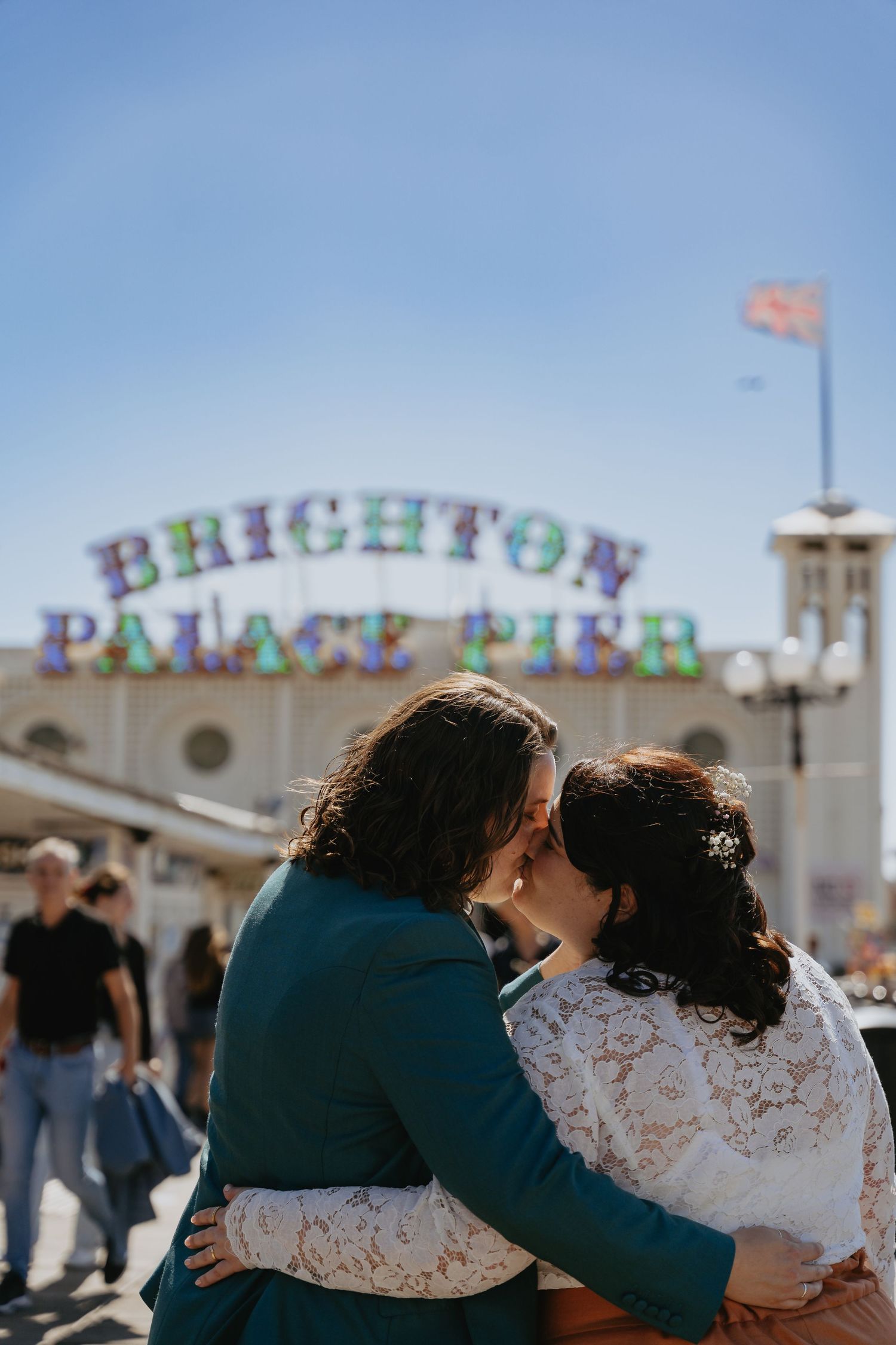 Two brides share a romantic kiss in front of the iconic Brighton Palace Pier sign on a sunny day.