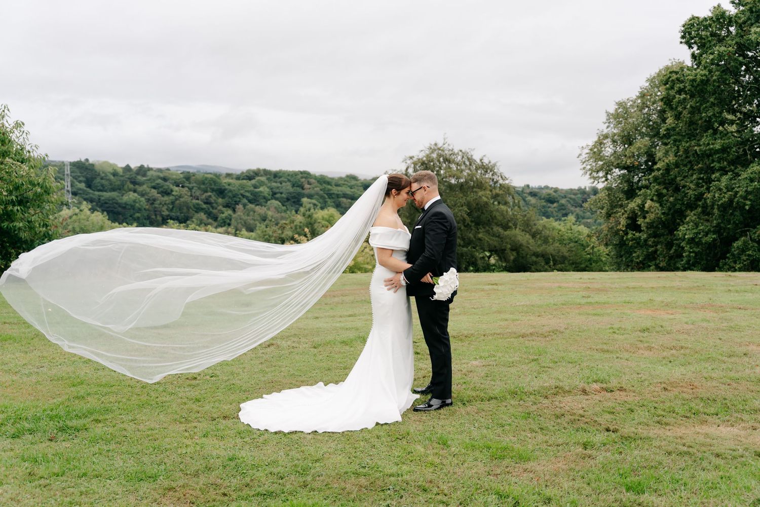 Bride and groom share a romantic kiss outdoors, with a flowing cathedral veil billowing in the wind on a lush green lawn.