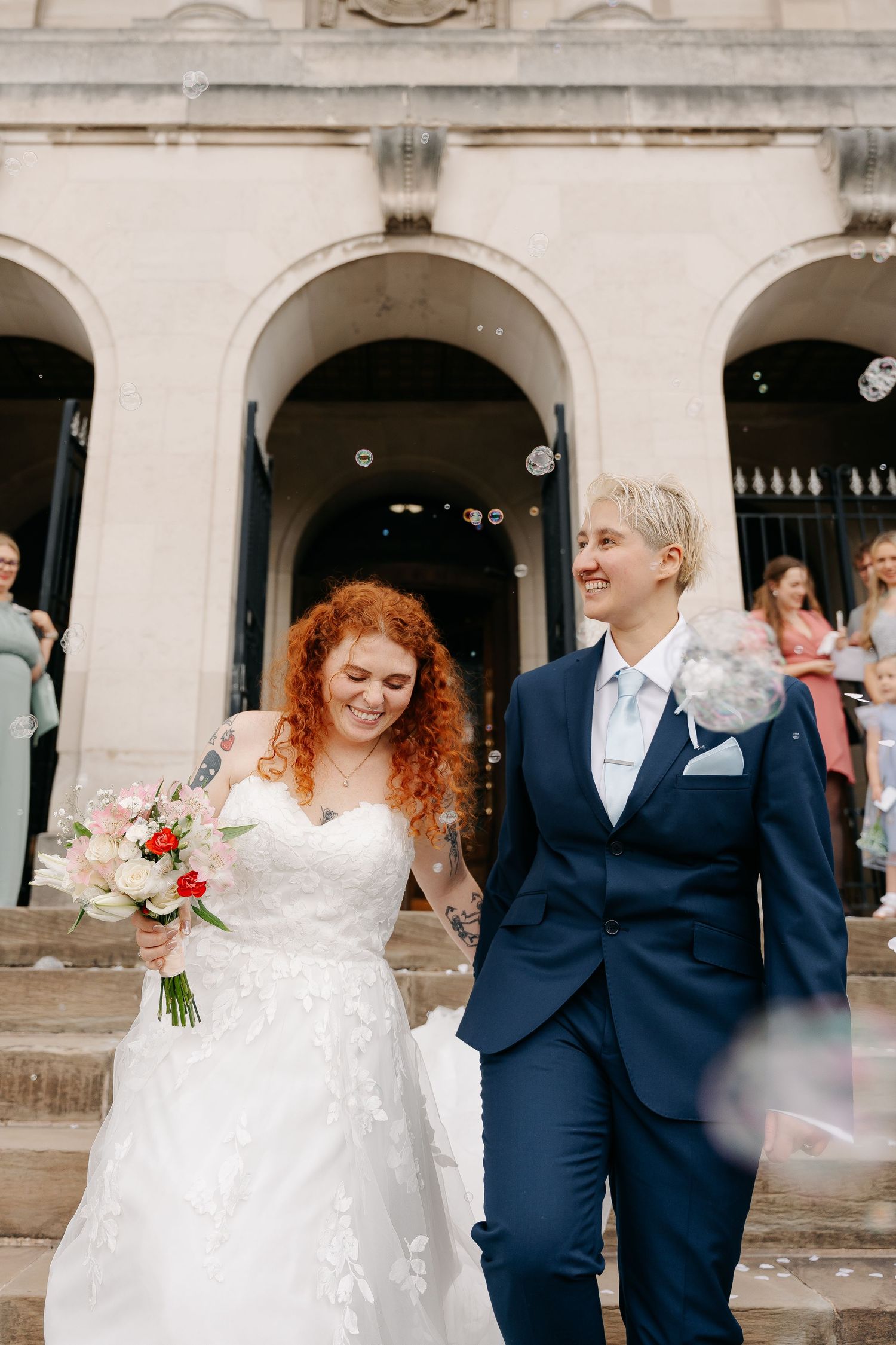 Two brides exit a grand building with bubbles, one in a white lace gown with bouquet, the other in a navy suit.
