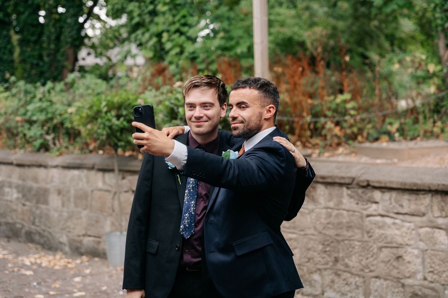 Two grooms in suits take a selfie outdoors near a stone wall, smiling on their wedding day.