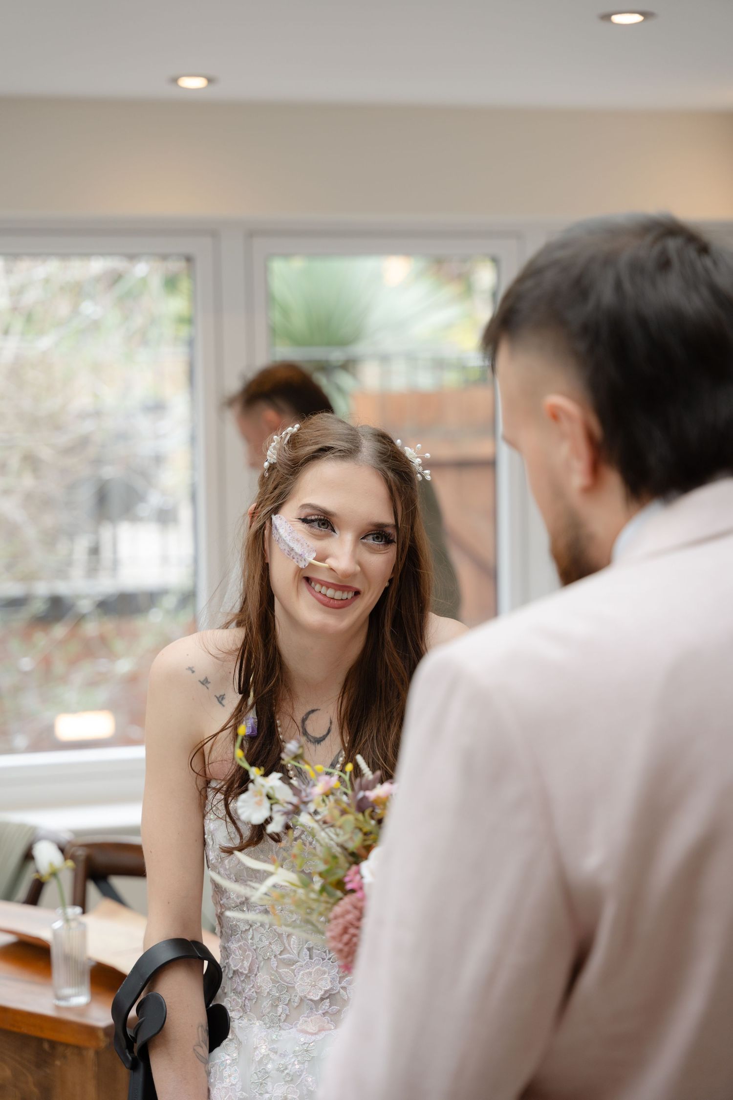 Smiling bride holding a wildflower bouquet gazes at groom during intimate indoor wedding ceremony.