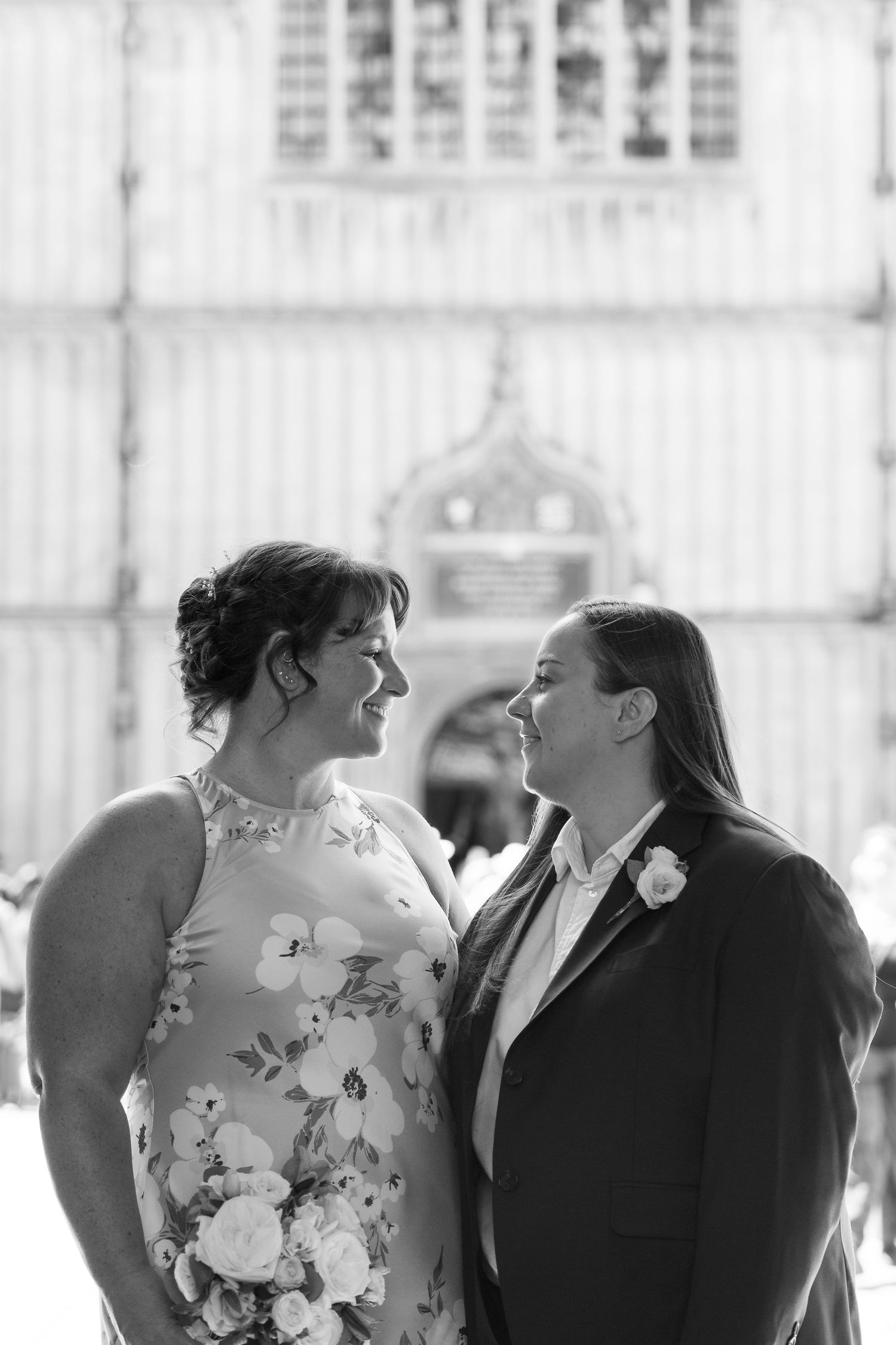 Black and white photo of two brides gazing at each other, one in floral dress, one in suit, outside a historic building.