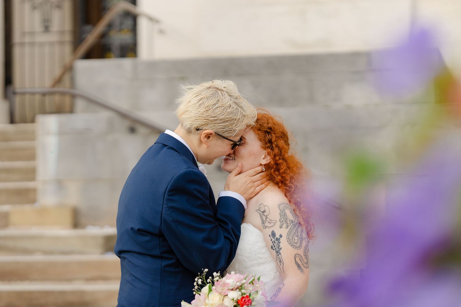 Two brides share a romantic kiss on wedding day, one in blue suit with blonde hair, other in white dress with red curly hair.