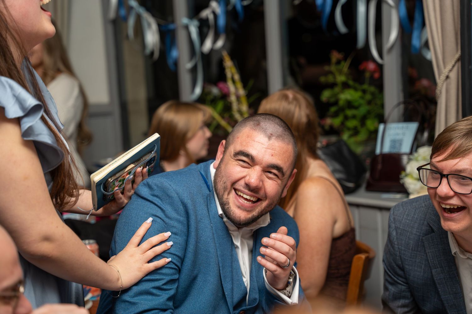 Man in blue suit laughing joyfully at a celebration event, surrounded by guests in a decorated venue.