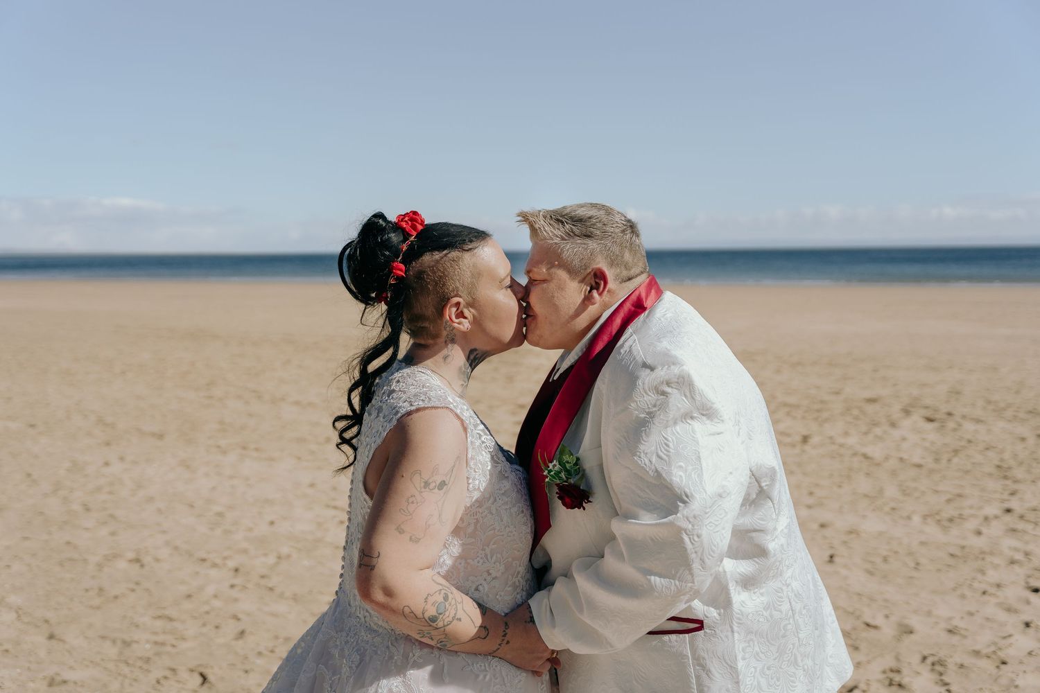 Couple sharing a romantic kiss on a sandy beach in wedding attire, with clear blue skies in the background.