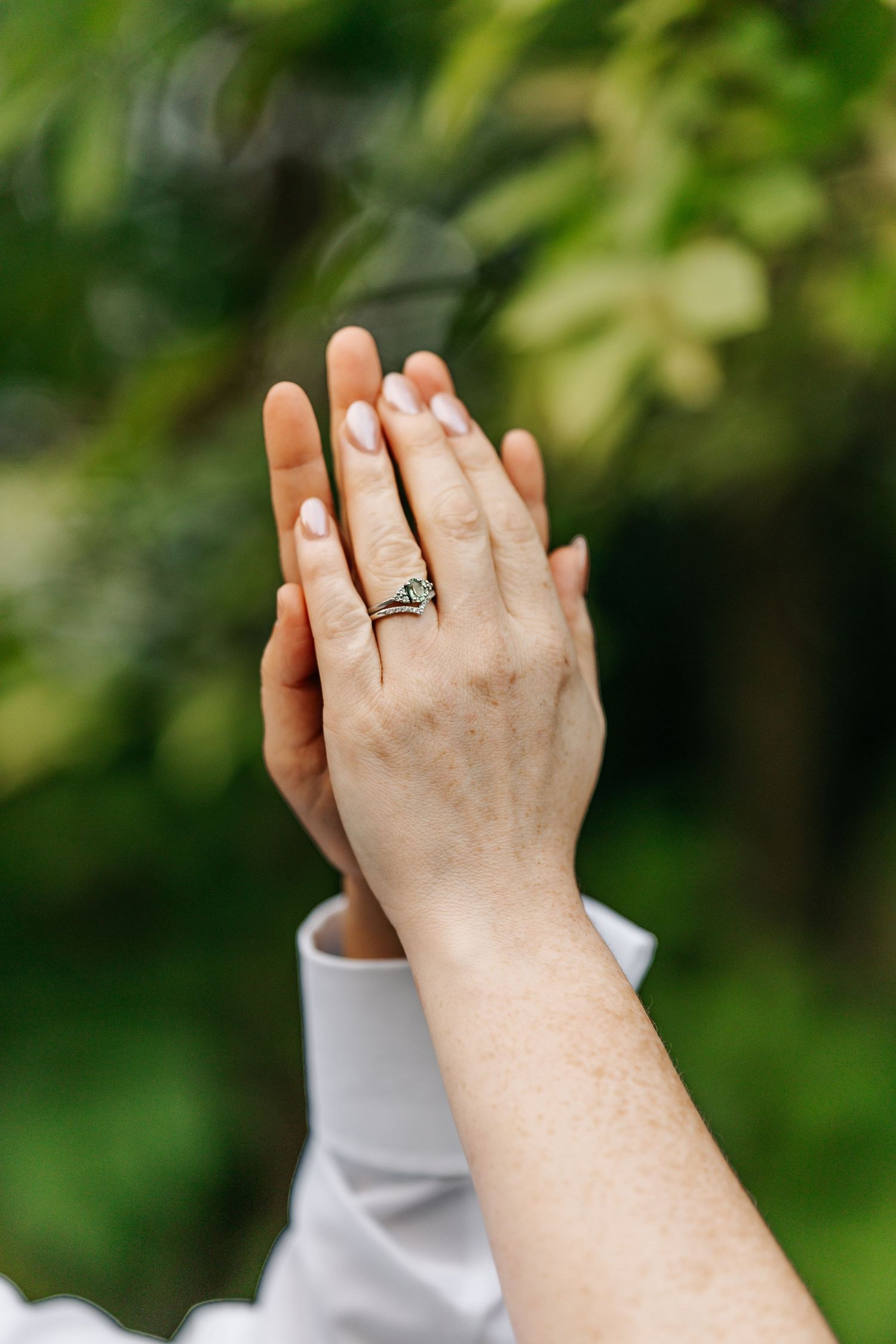 Two hands touching outdoors, one adorned with a delicate diamond engagement ring, set against a lush green garden background.