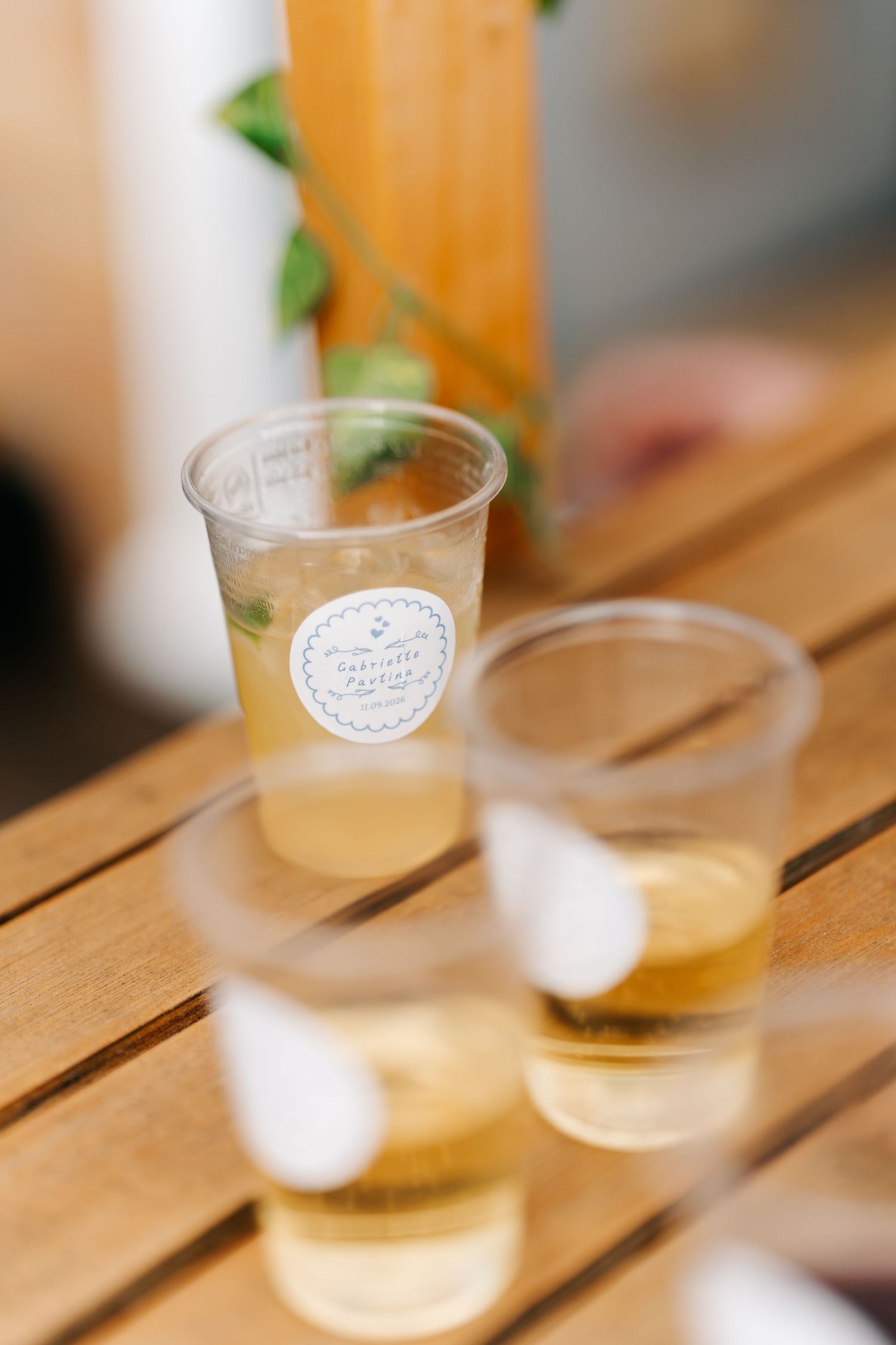 Three plastic cups with yellow drinks and a branded sticker sit on a wooden table at an outdoor event.