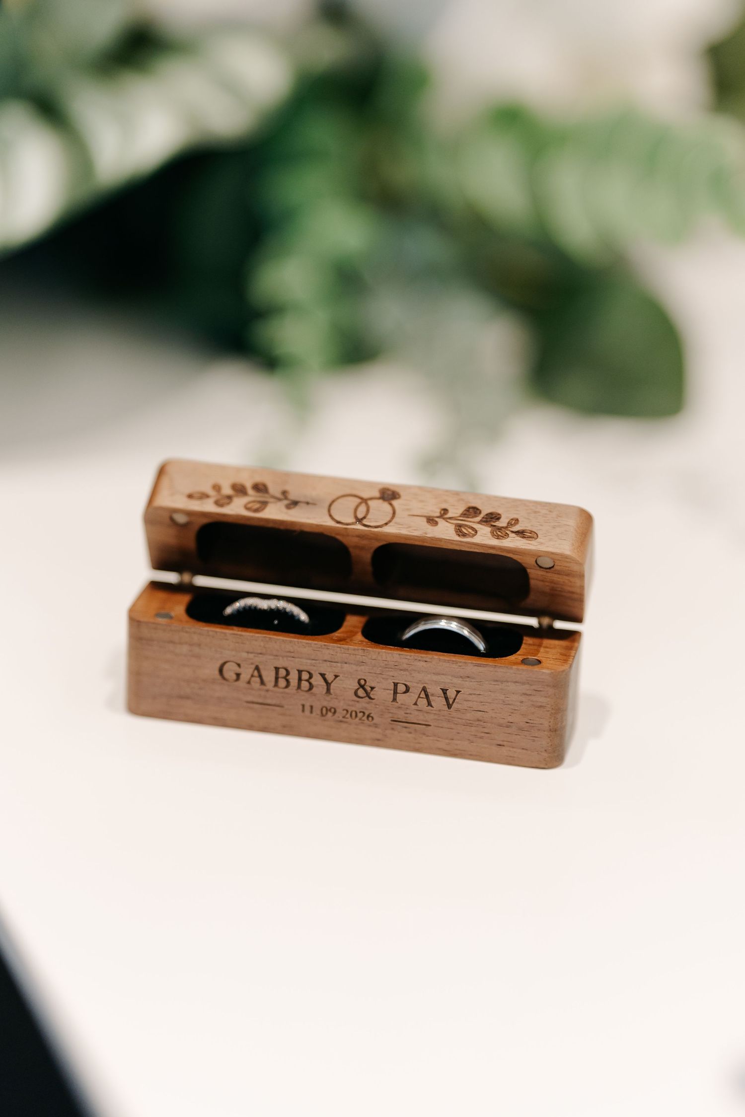 Personalized wooden wedding ring box engraved with Gabby & Pav holding two wedding bands, with floral ring design on lid.
