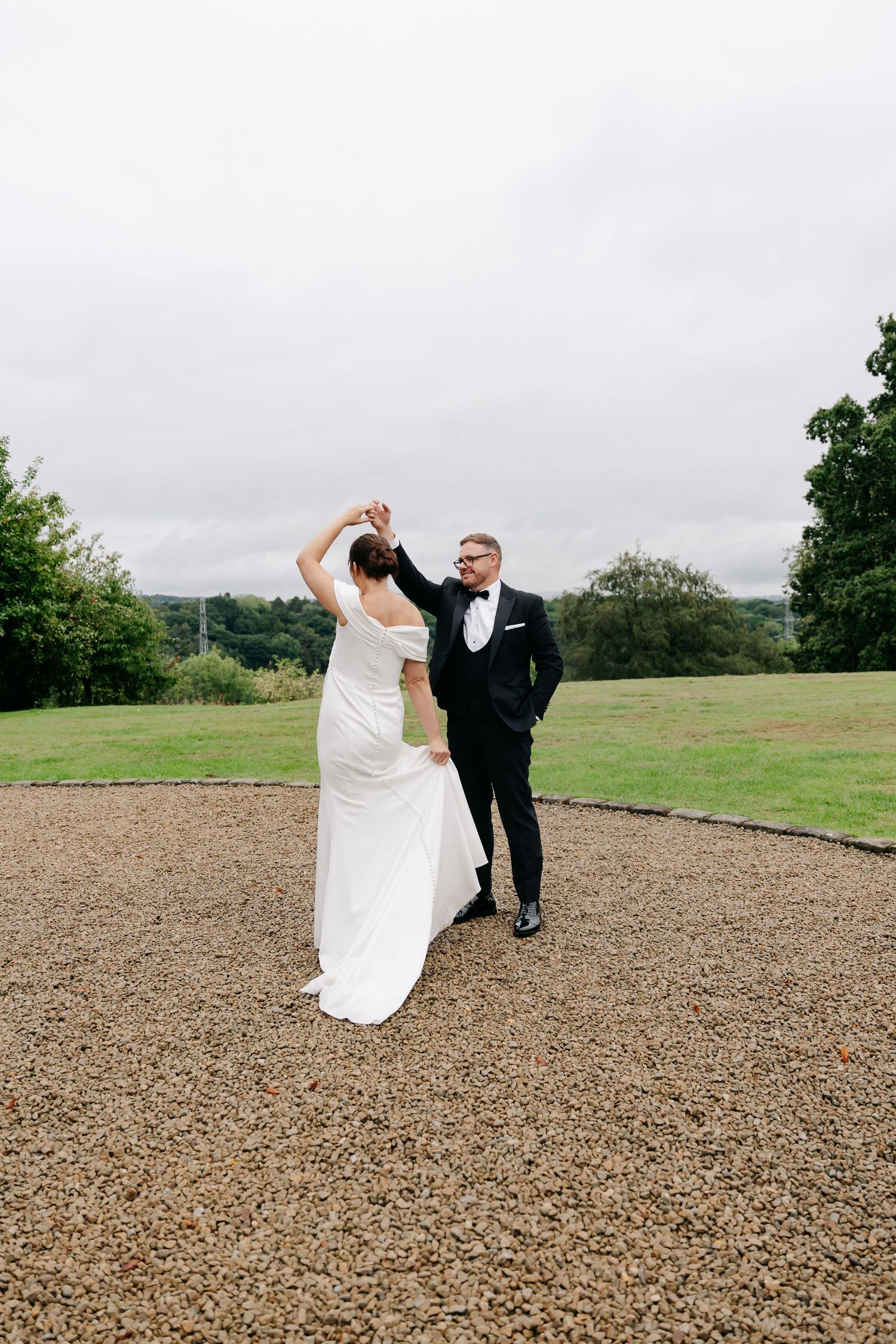 Bride and groom dancing outdoors on gravel path, lush green lawn and trees in background on their wedding day.