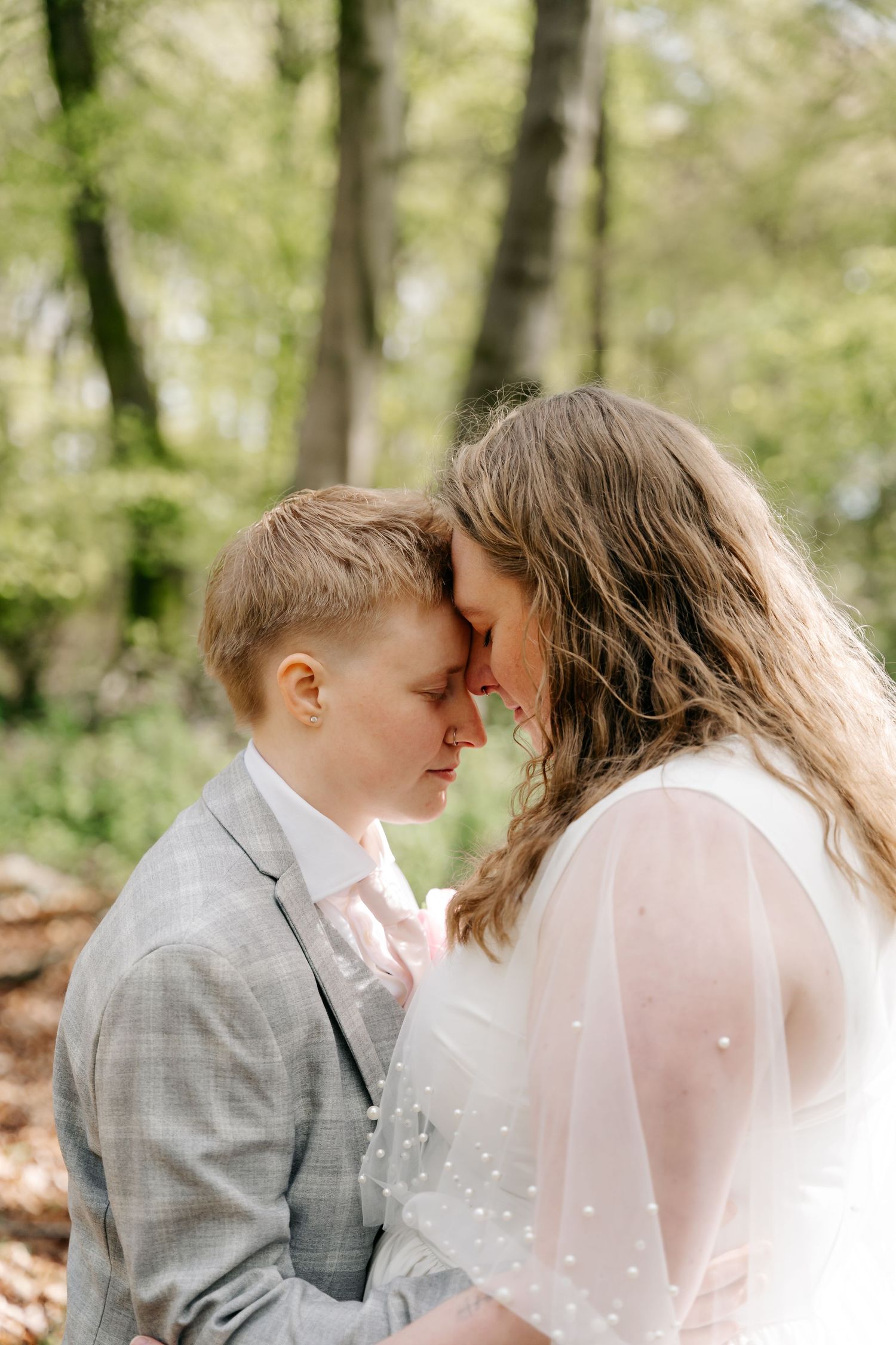 Two brides share an intimate forehead touch moment outdoors, one in a gray suit and one in a white wedding dress.