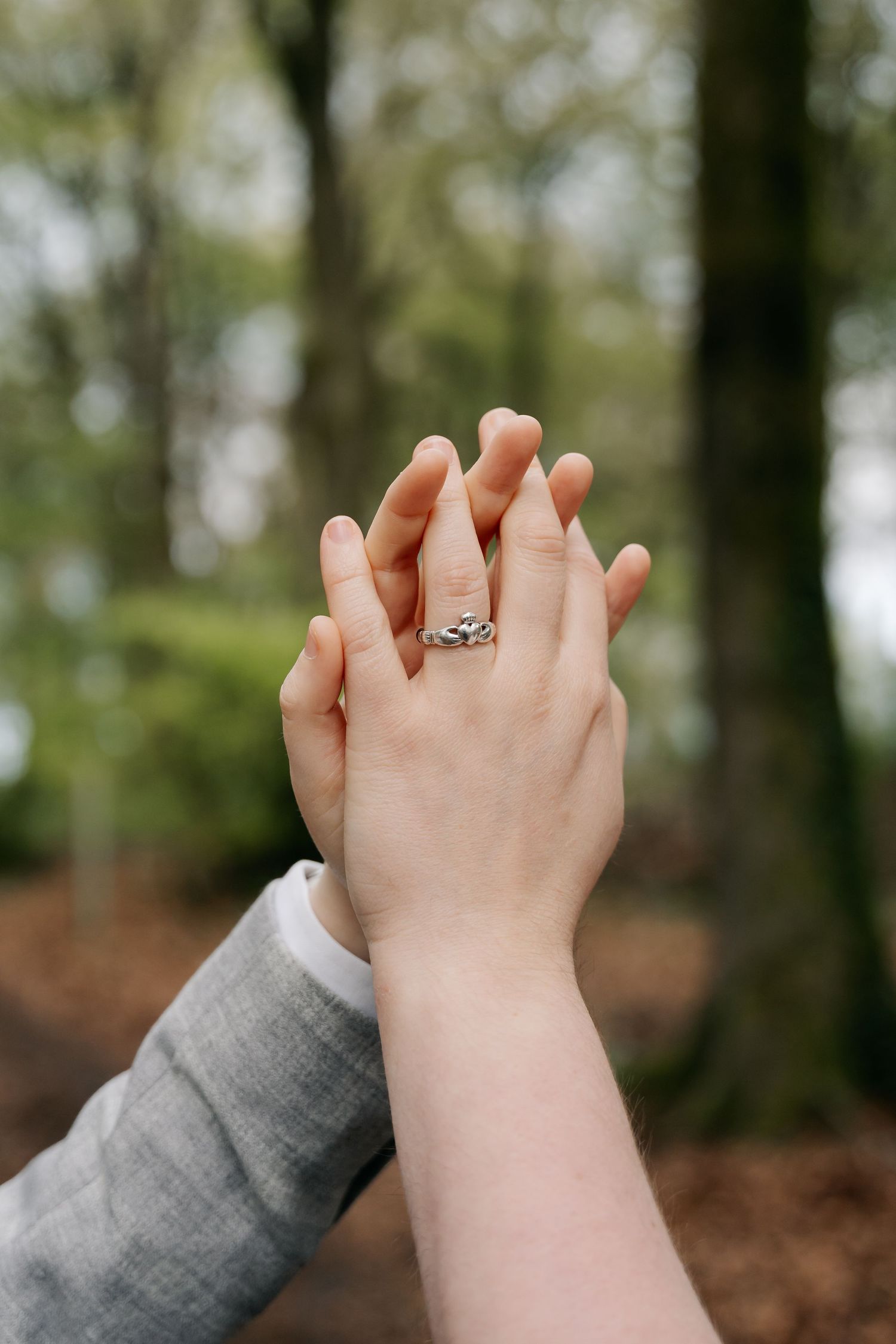 Two hands intertwined in a forest, one wearing a silver claddagh ring, symbolizing love and commitment.