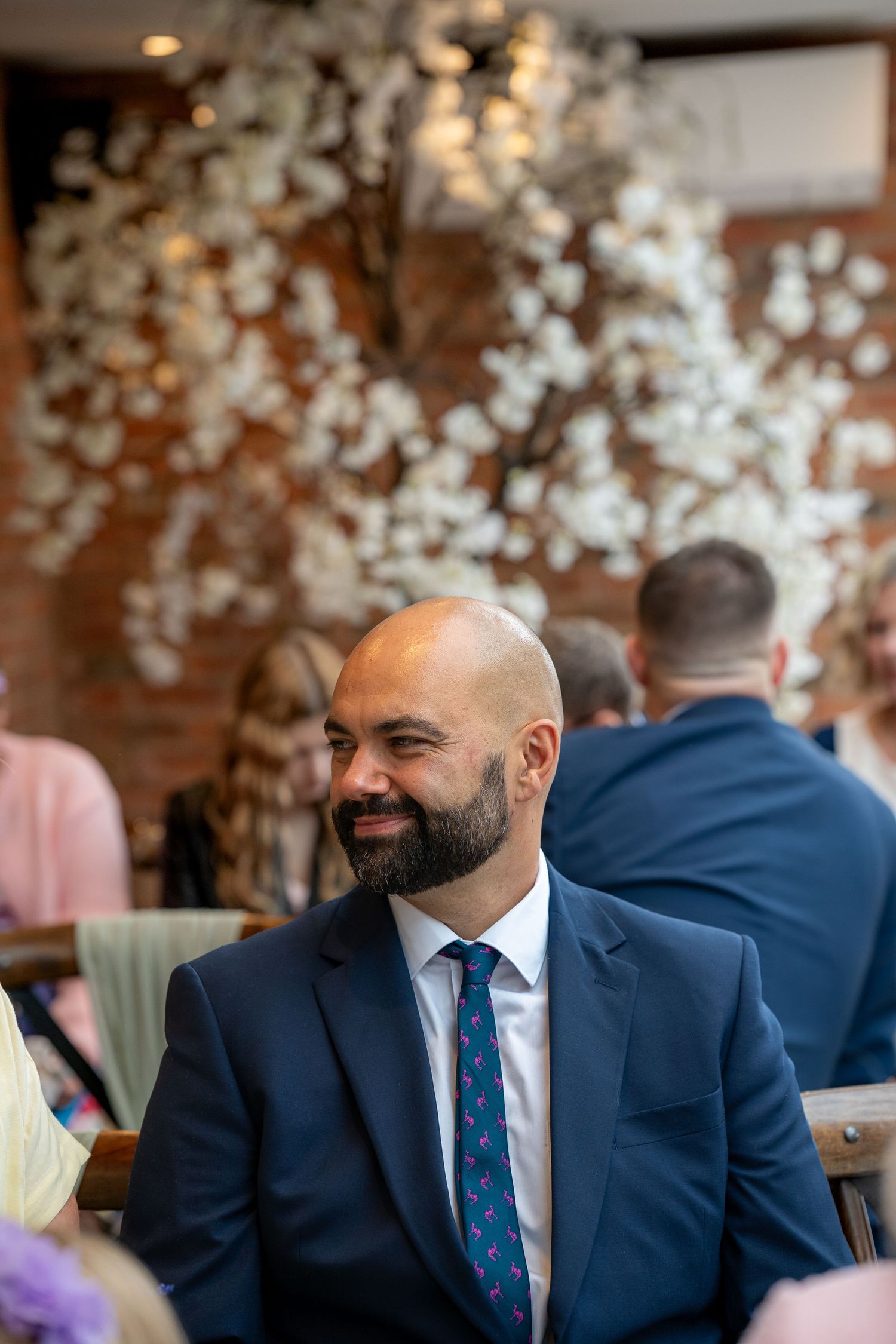 Smiling bald man with beard in navy suit and patterned tie seated at an elegant wedding venue with white floral decor.