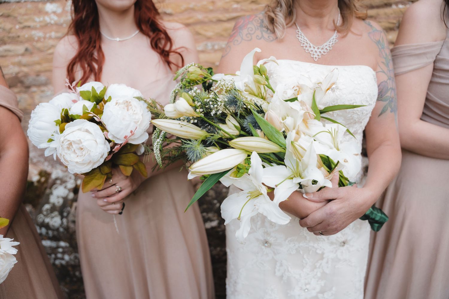 Bride holding white lily bouquet flanked by bridesmaids in taupe dresses with white peony bouquets at outdoor wedding.