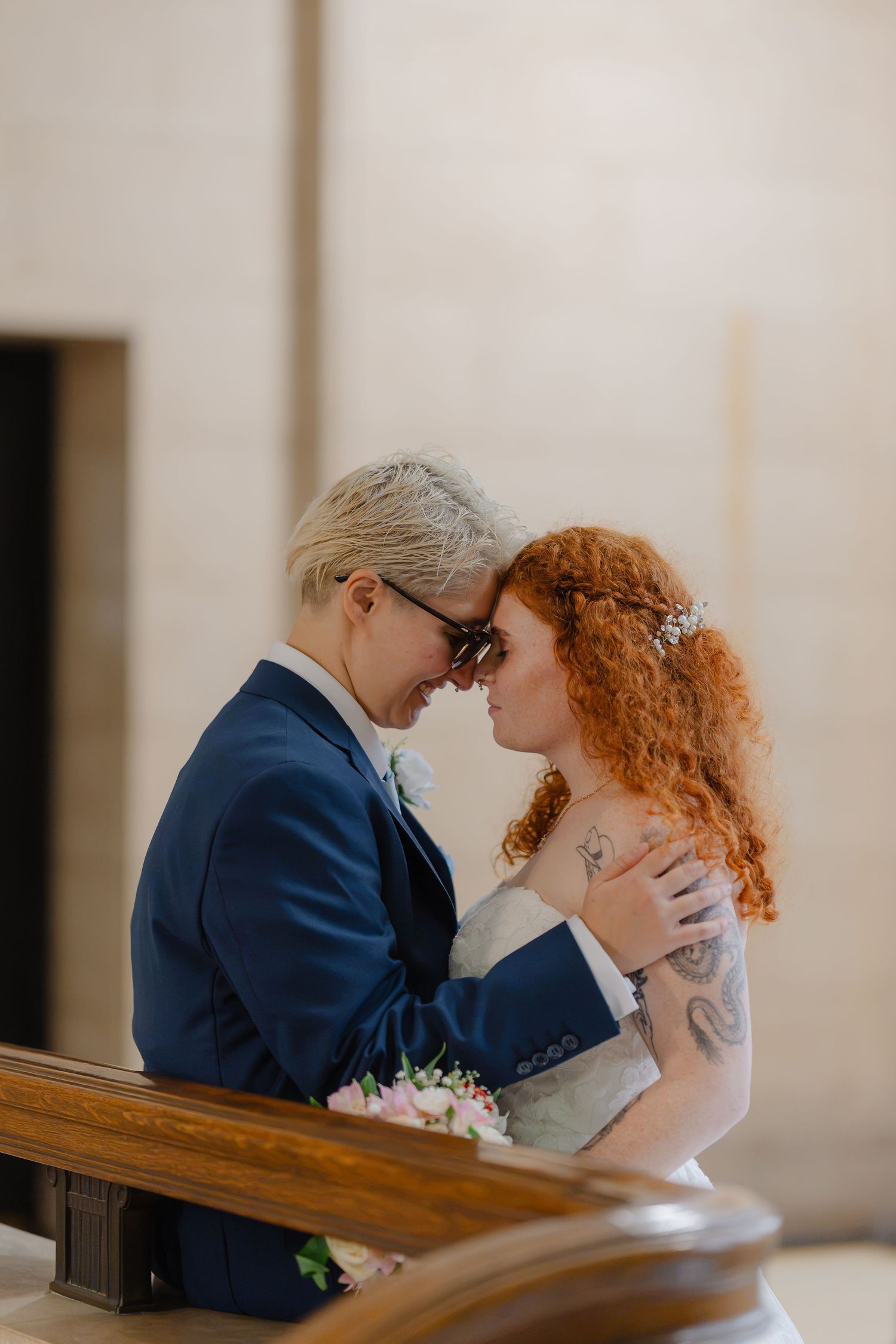 Two brides share an intimate moment, one in a blue suit and one in a white gown, touching foreheads at their wedding.