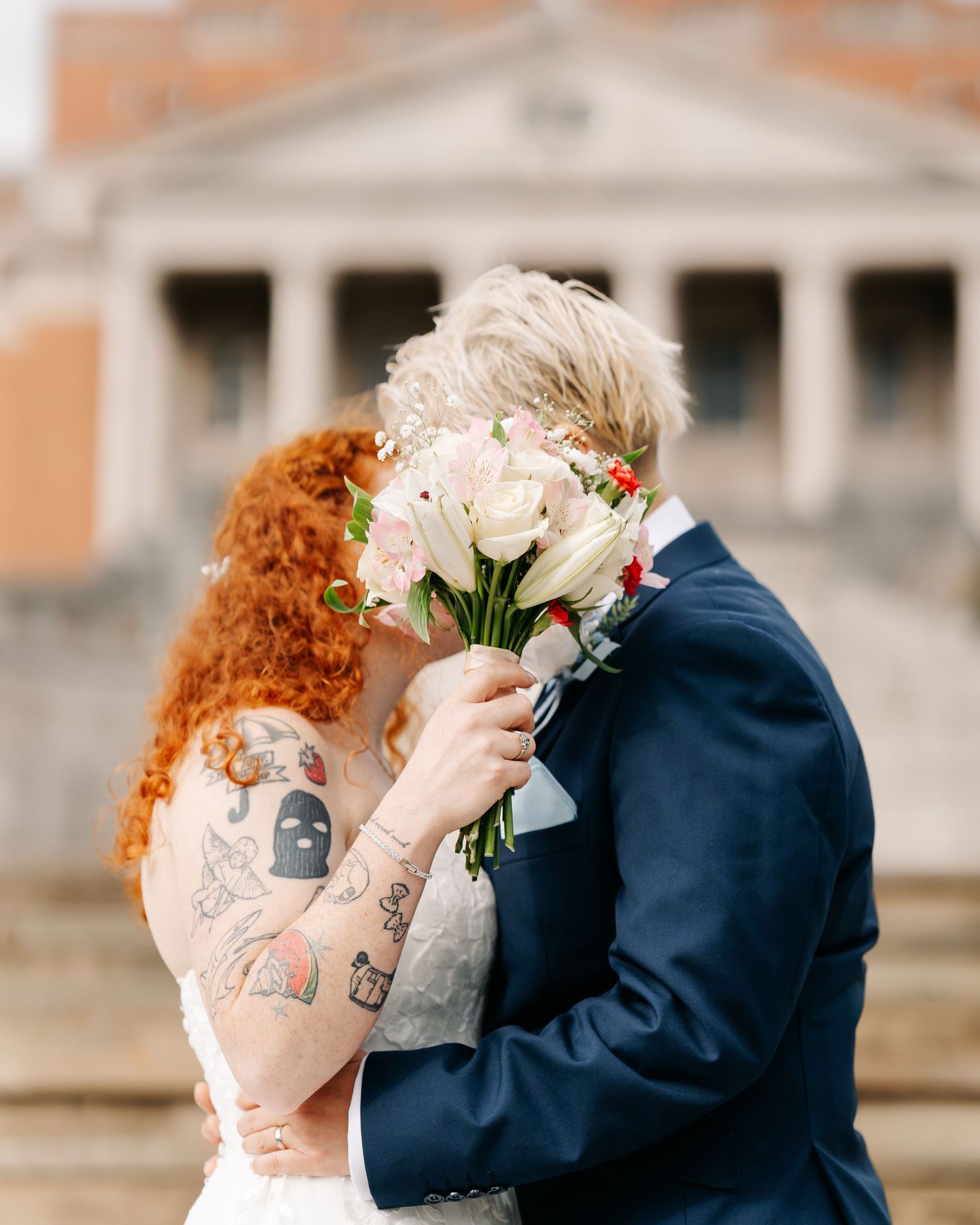 Tattooed bride with red hair kisses groom in navy suit, bouquet of pink and white flowers hiding their faces.