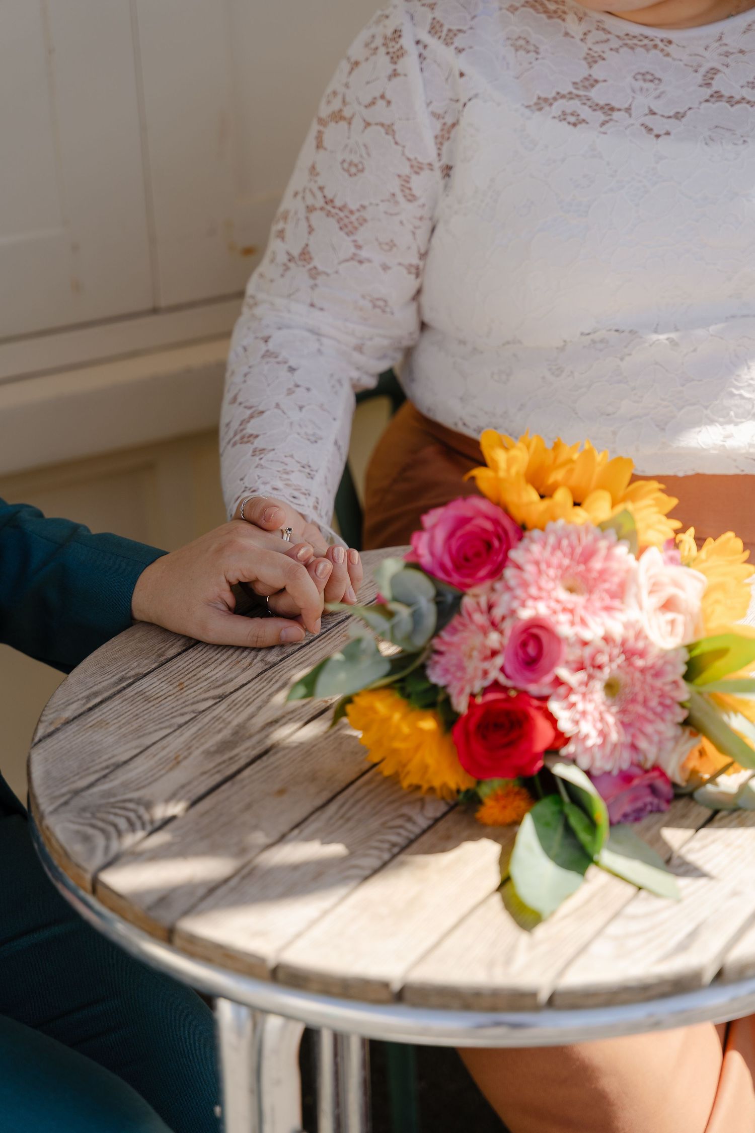 Couple holding hands at a wooden table with a colorful bridal bouquet of pink, red, and yellow flowers.