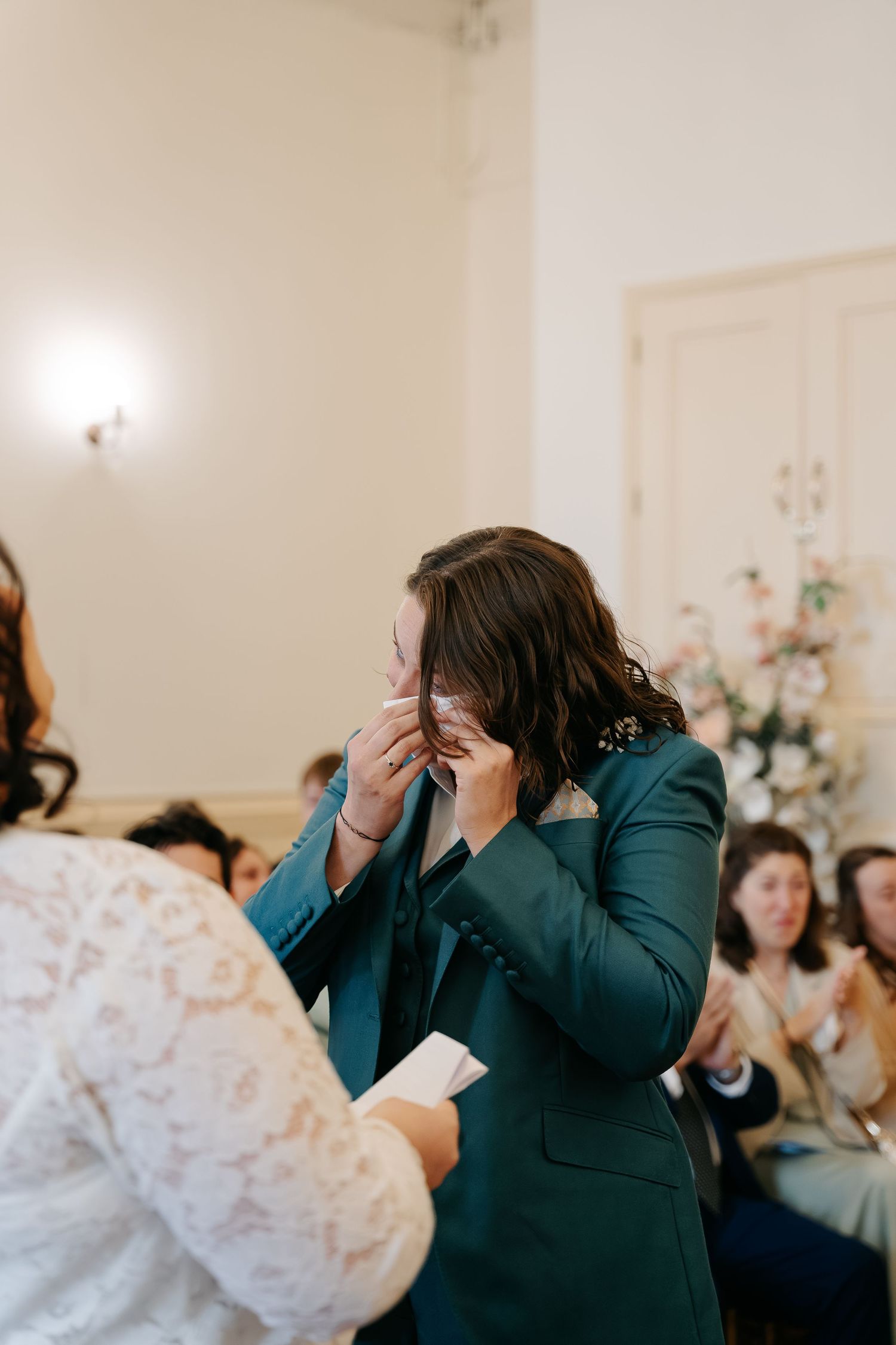 Emotional partner in teal suit wiping tears during wedding ceremony while guests applaud in elegant venue.