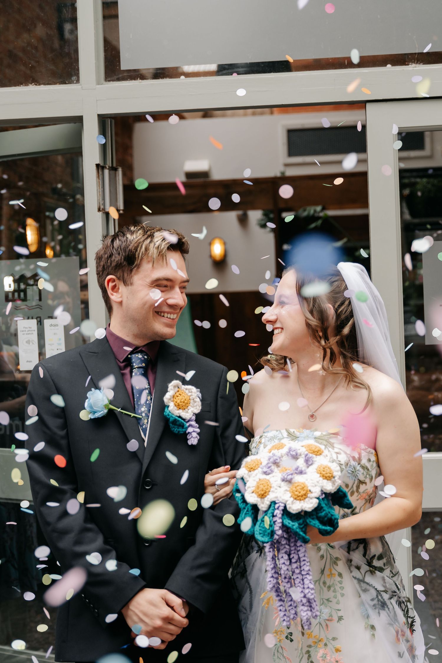 Newlyweds smile at each other as colorful confetti falls around them outside a venue, bride holding crochet flower bouquet.