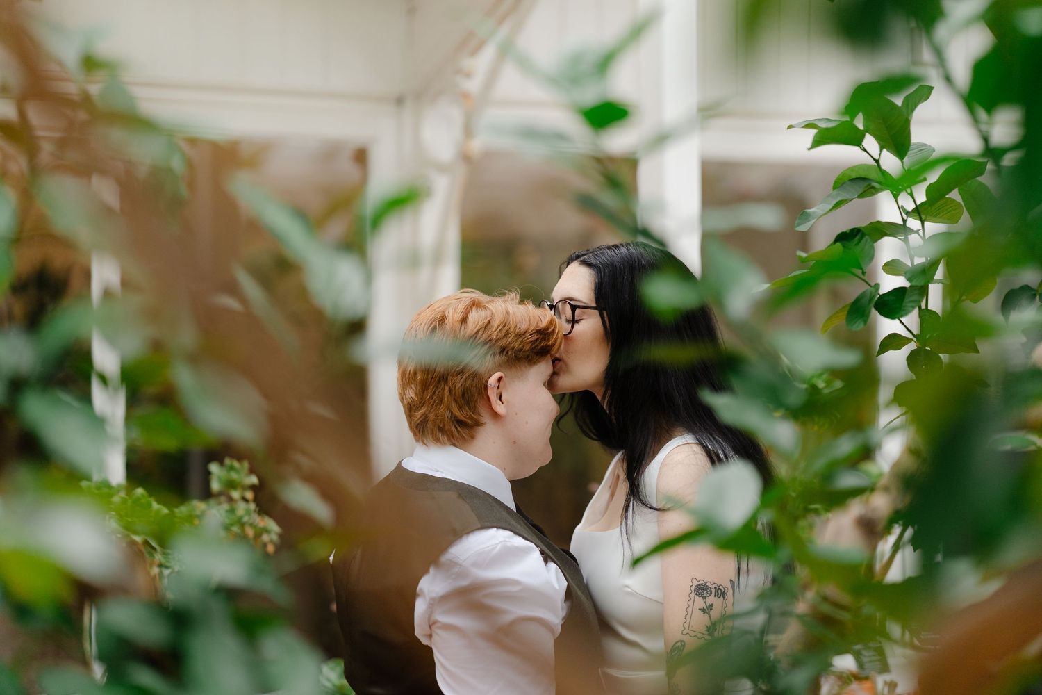 Couple sharing a romantic kiss surrounded by lush green plants in a greenhouse wedding setting.