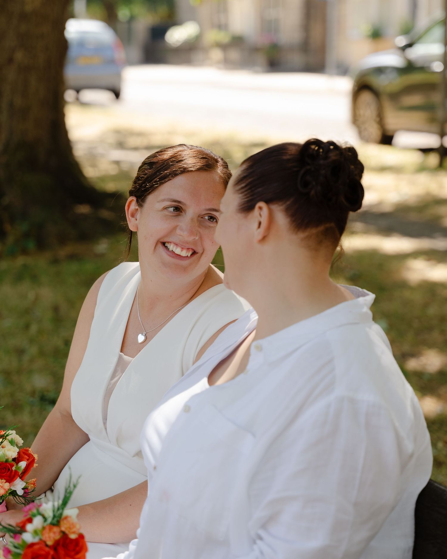 Two brides in white attire share a joyful moment outdoors, one smiling brightly while holding a colorful bouquet.