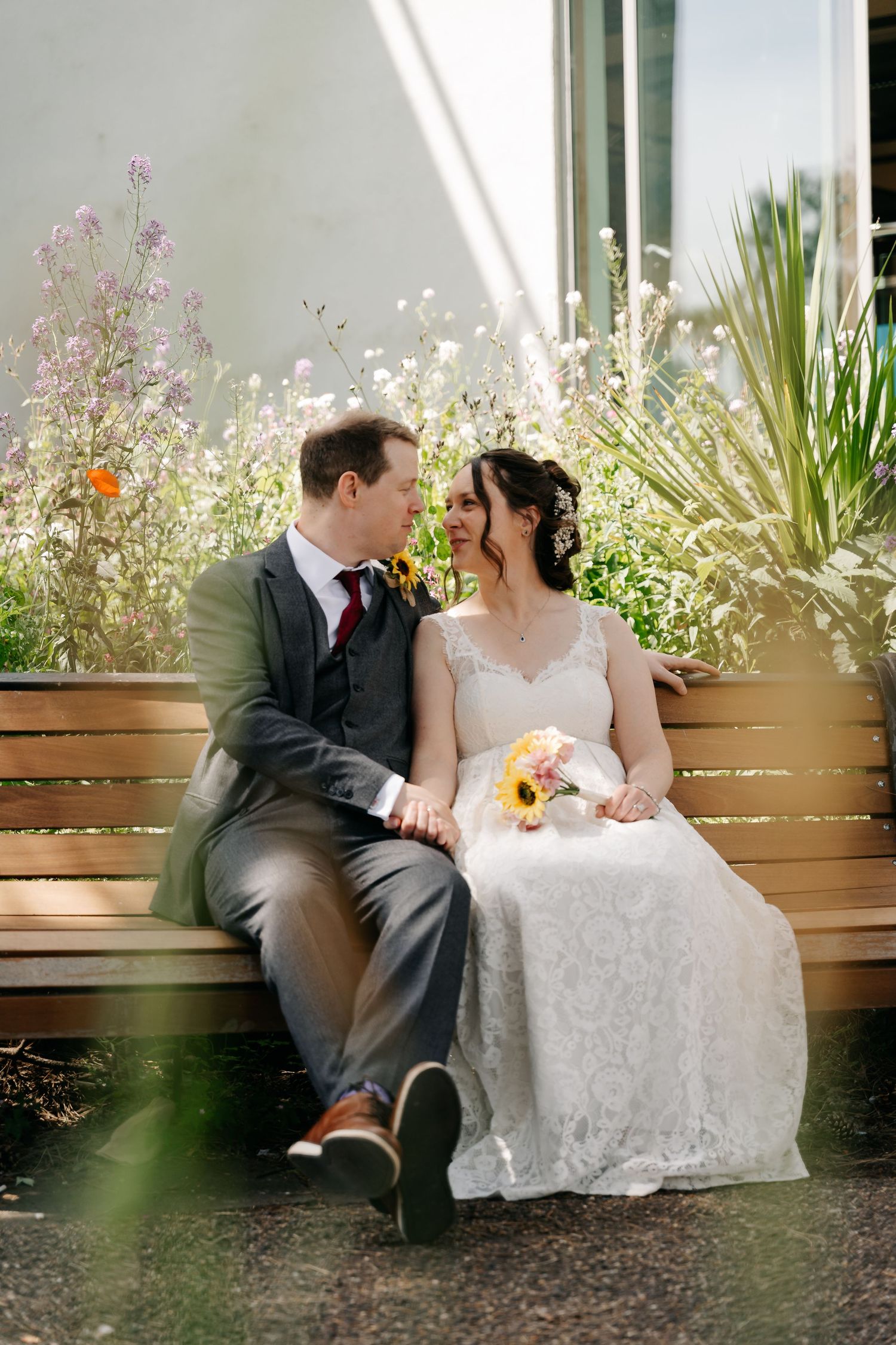 A bride and groom sit together on a wooden bench surrounded by wildflowers, sharing a tender moment on their wedding day.