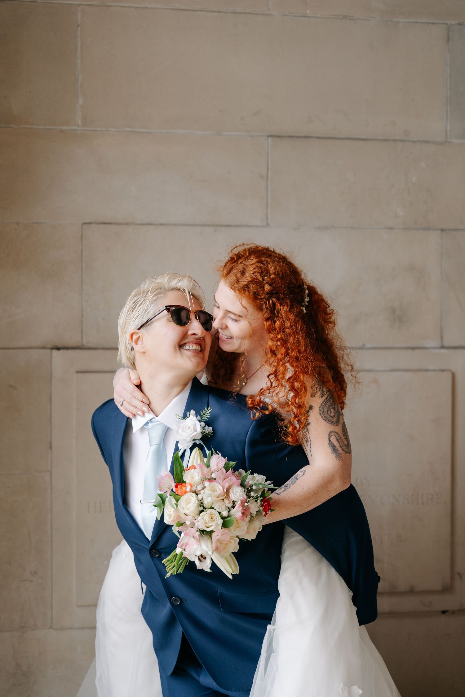 Two brides in wedding attire embrace and smile, one in a navy suit with bouquet, the other in a white gown with red curly hair.
