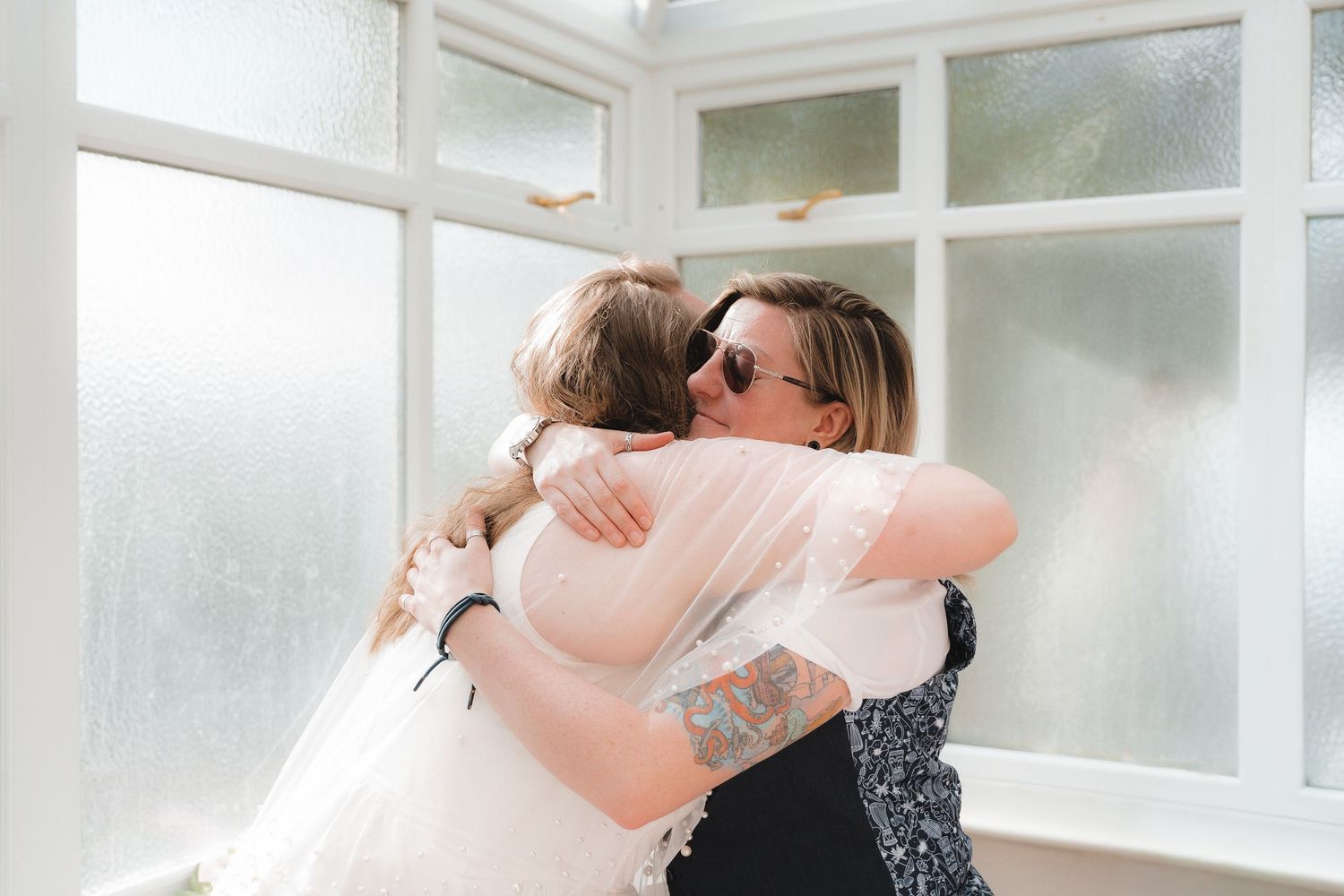 Bride in white gown and veil sharing a warm hug with a tattooed guest in sunglasses near bright windows.