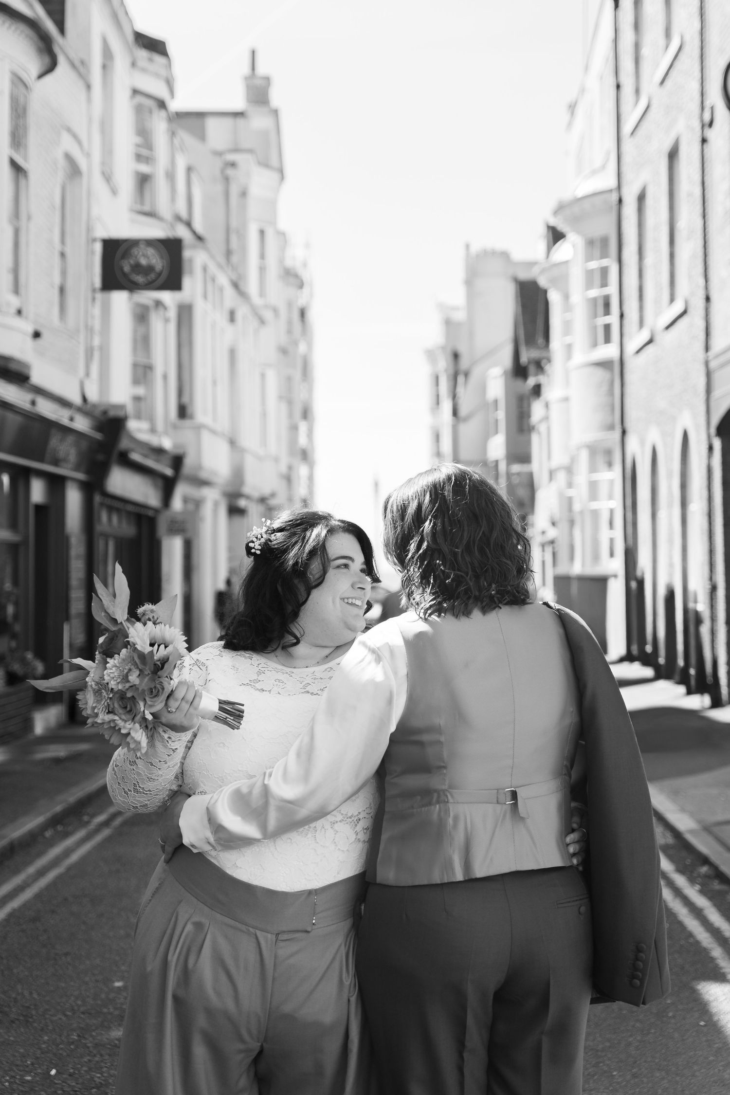 Black and white photo of a smiling bride holding flowers, walking arm in arm with partner down a sunny urban street.