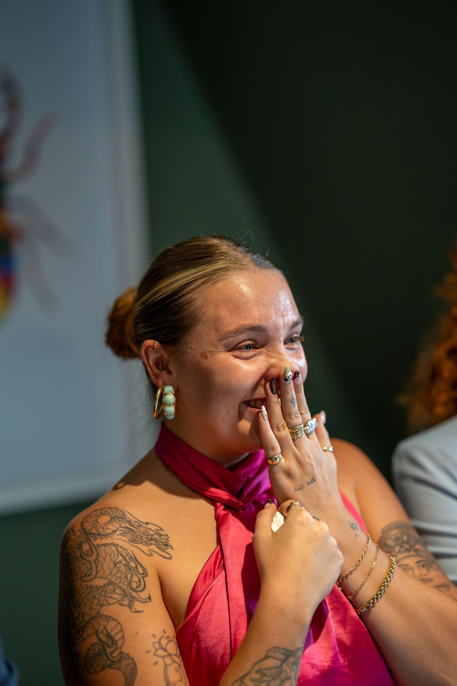 Laughing woman in pink halter top with tattoos and gold rings covering her mouth at an event.