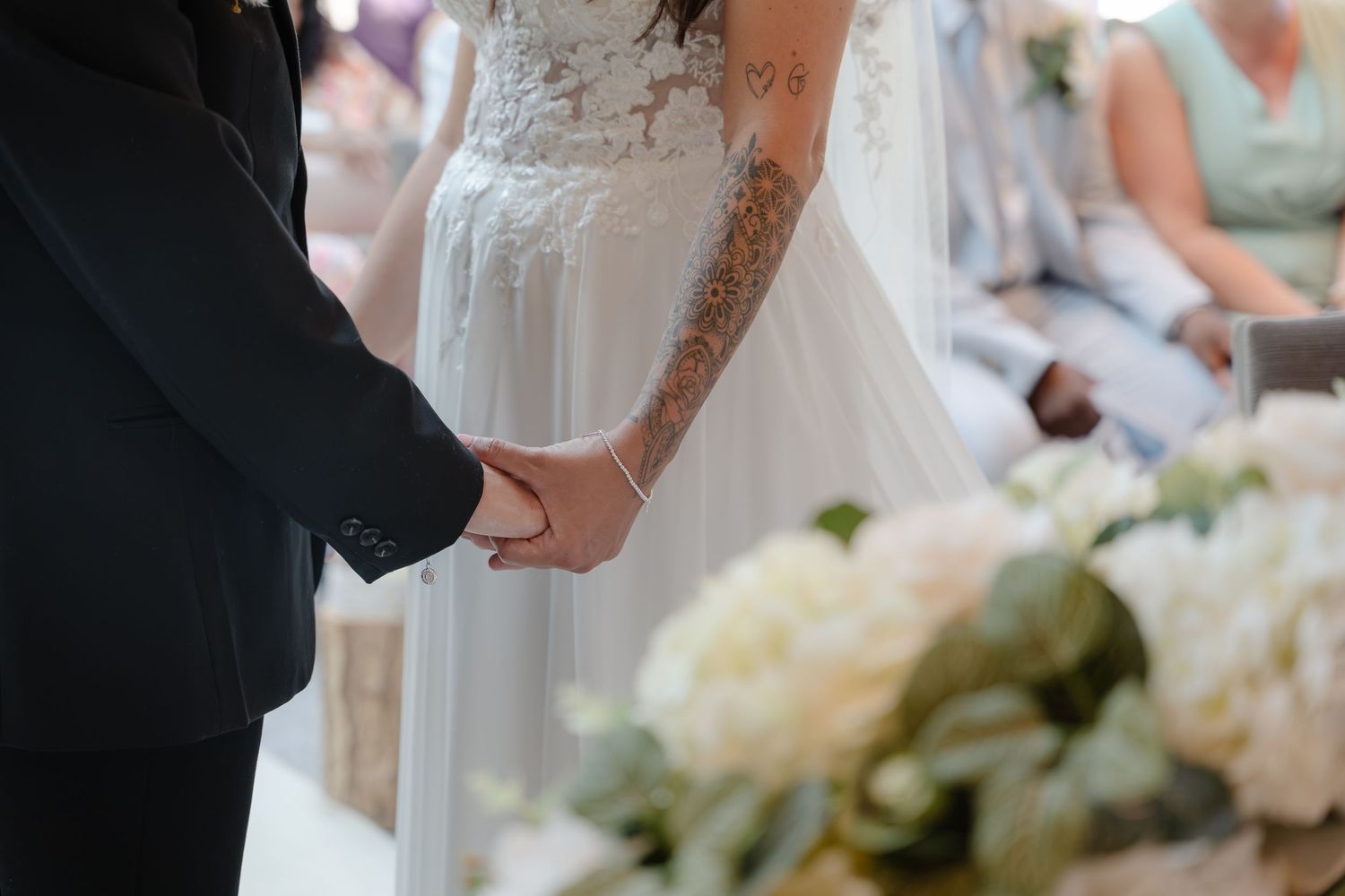 Bride and groom holding hands at wedding ceremony with white floral bouquet in foreground.