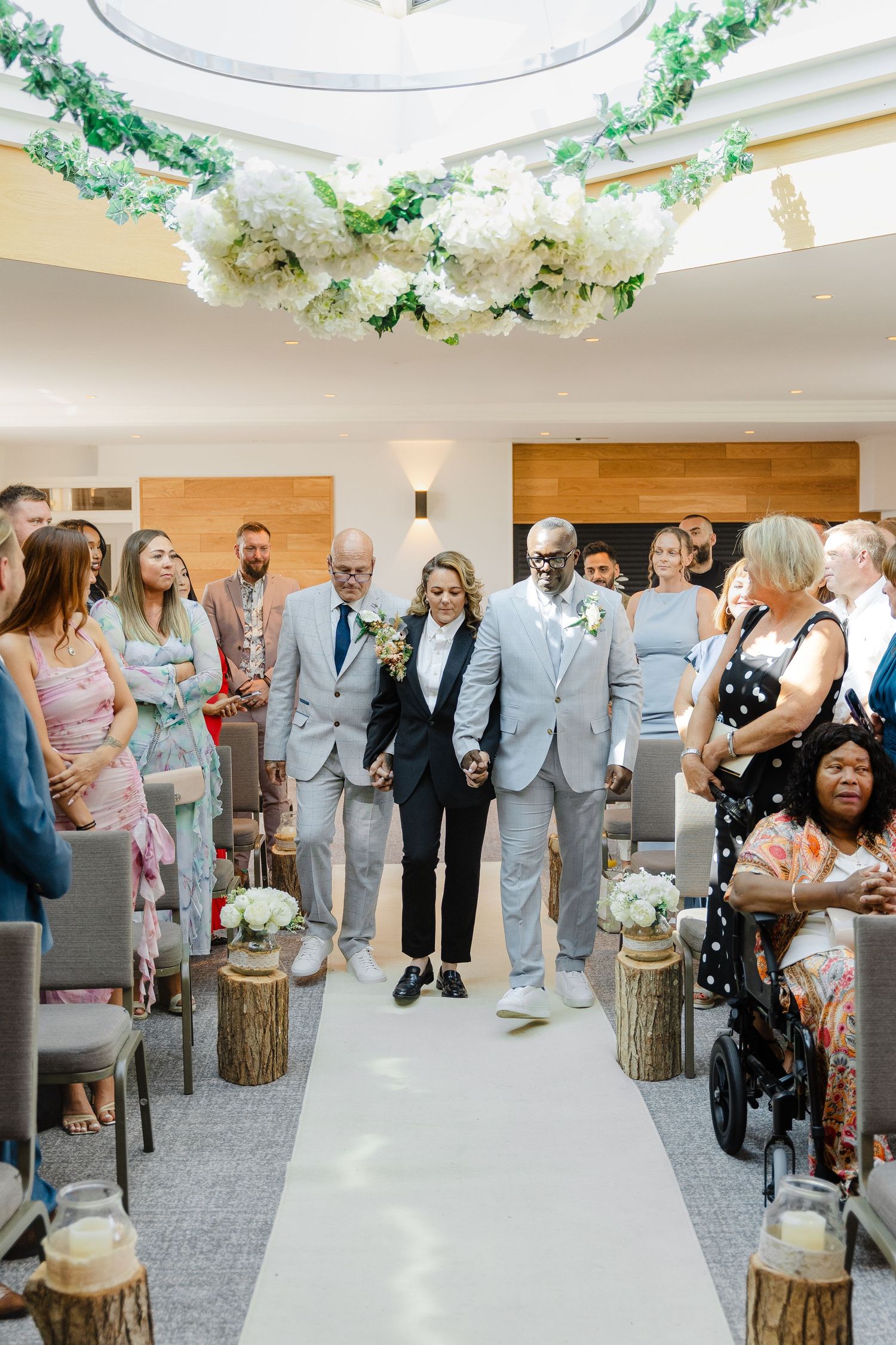 Bride walks down the aisle flanked by two escorts at an elegant indoor wedding ceremony with floral ceiling decor.