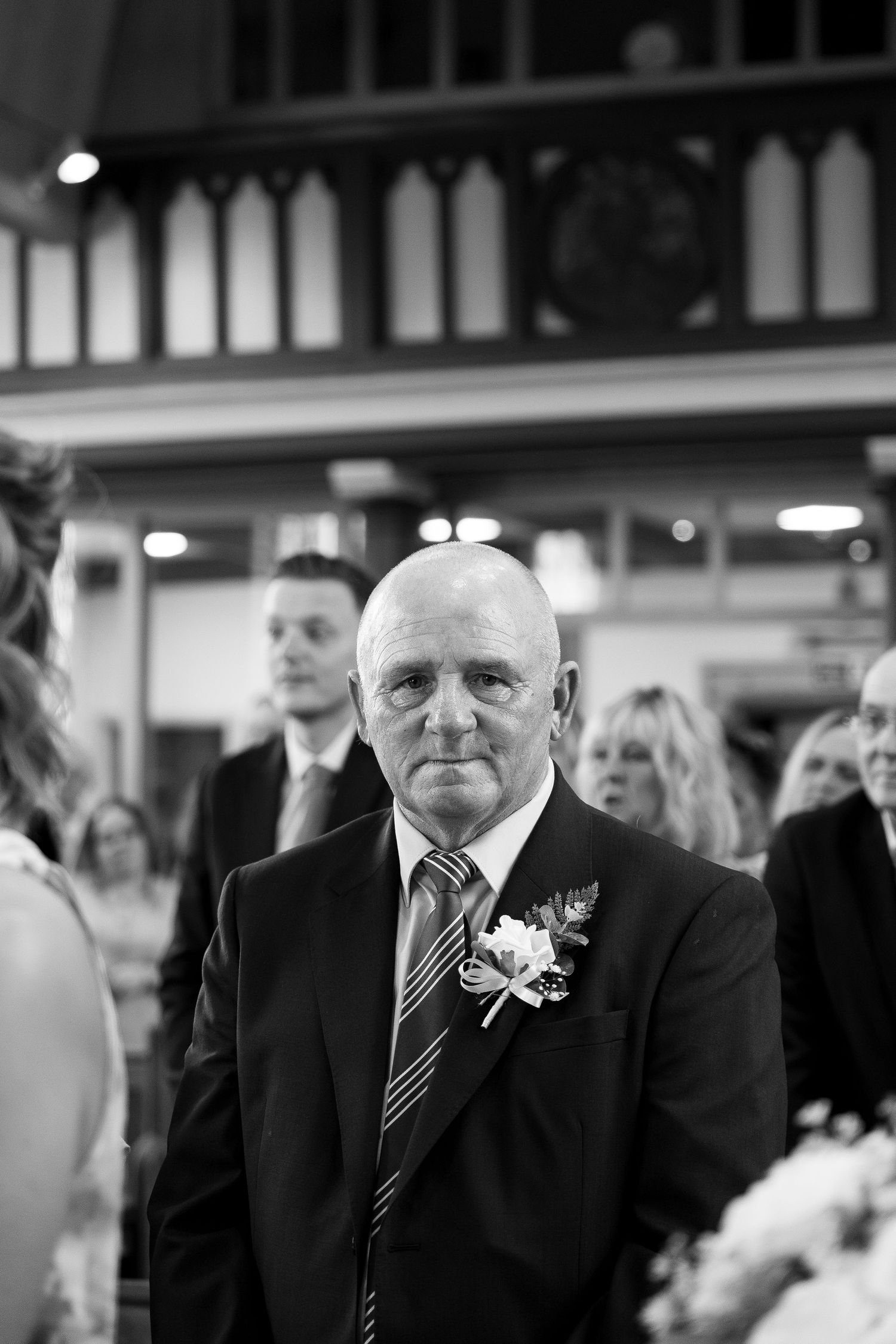 Black and white photo of a bald man in a suit with boutonniere at a wedding ceremony.