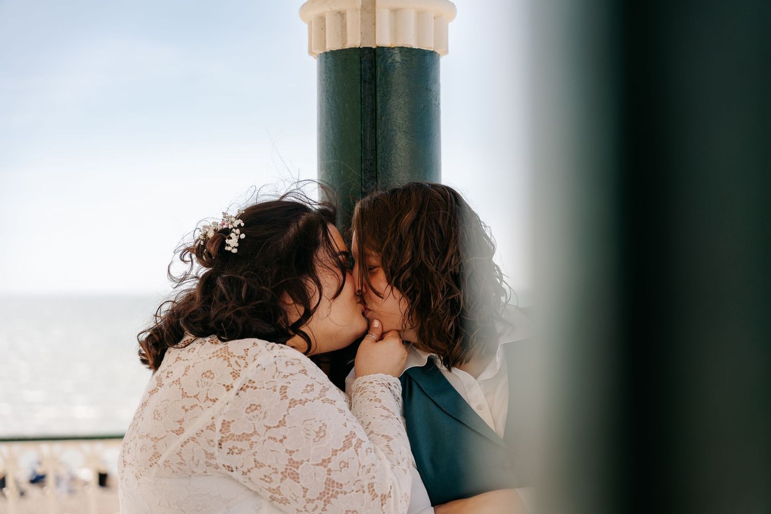 Two brides share a romantic kiss outdoors near a teal pillar with ocean views on their wedding day.