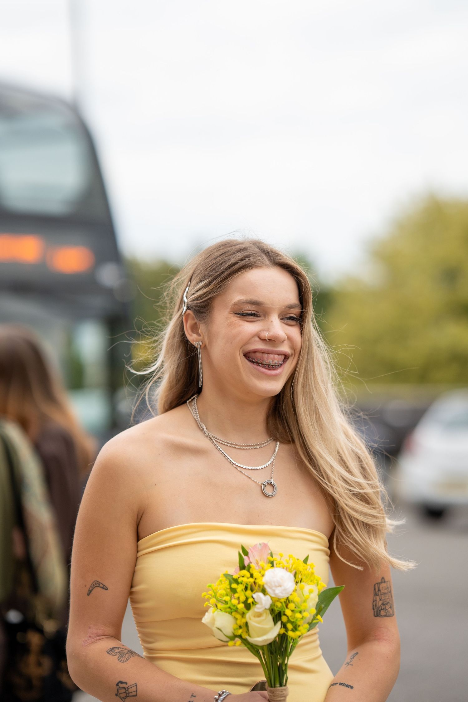 Young woman in yellow strapless dress with tattoos smiling and holding yellow and white flower bouquet outdoors.