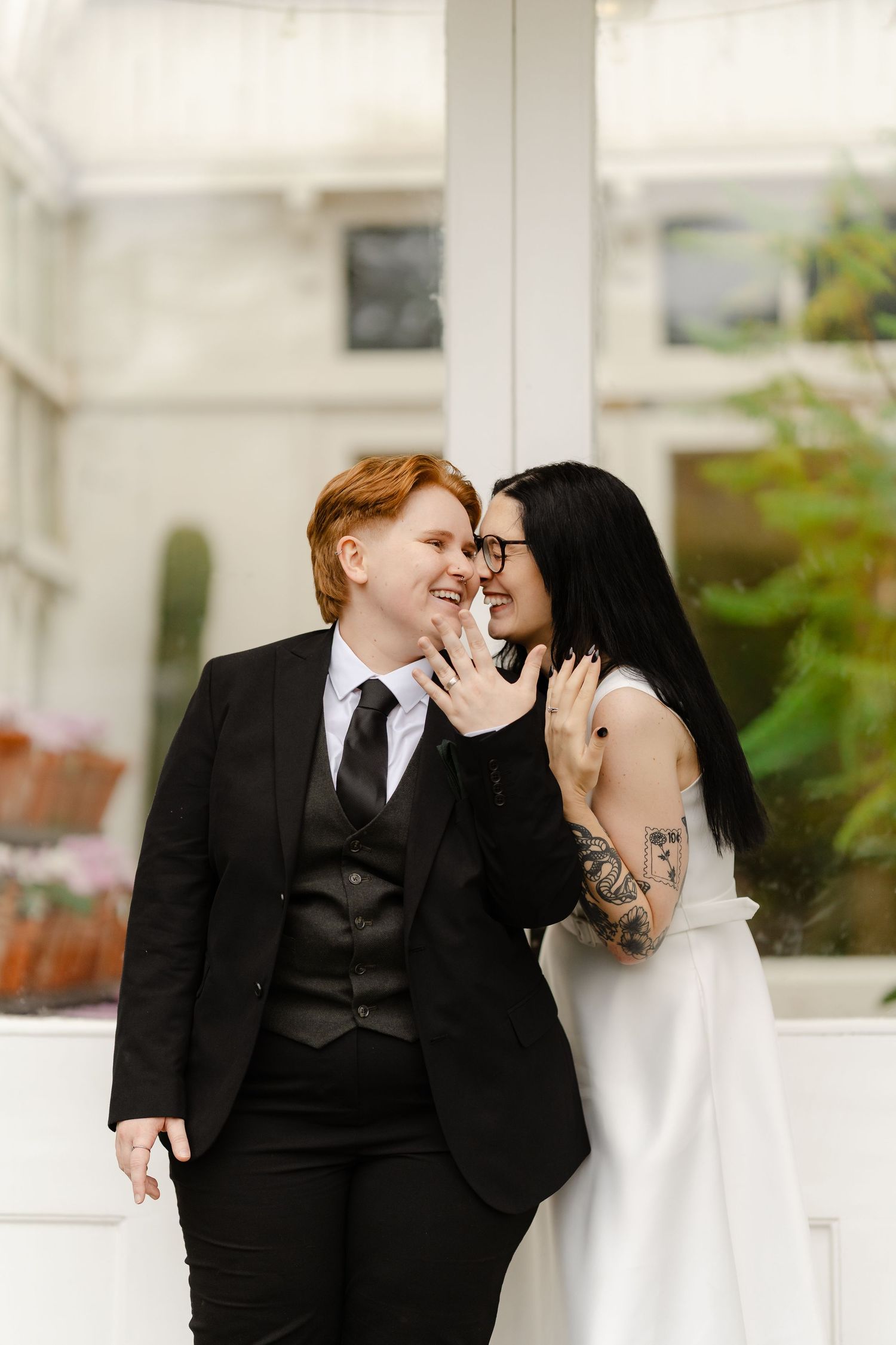 Two brides share a romantic moment, one in a black suit and one in a white dress with tattoos, at a wedding.