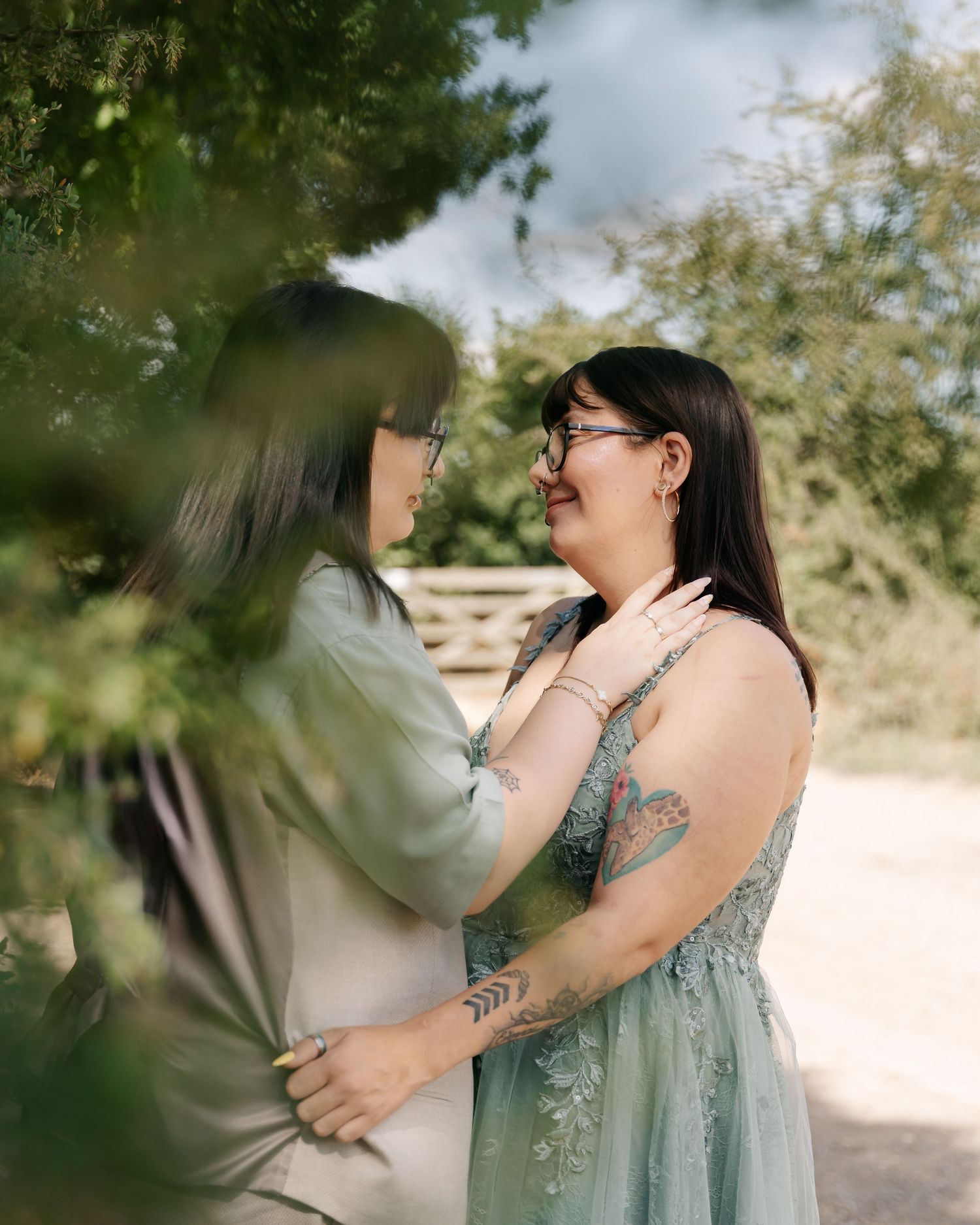 Two women in sage green outfits share a tender moment outdoors, framed by lush green foliage.